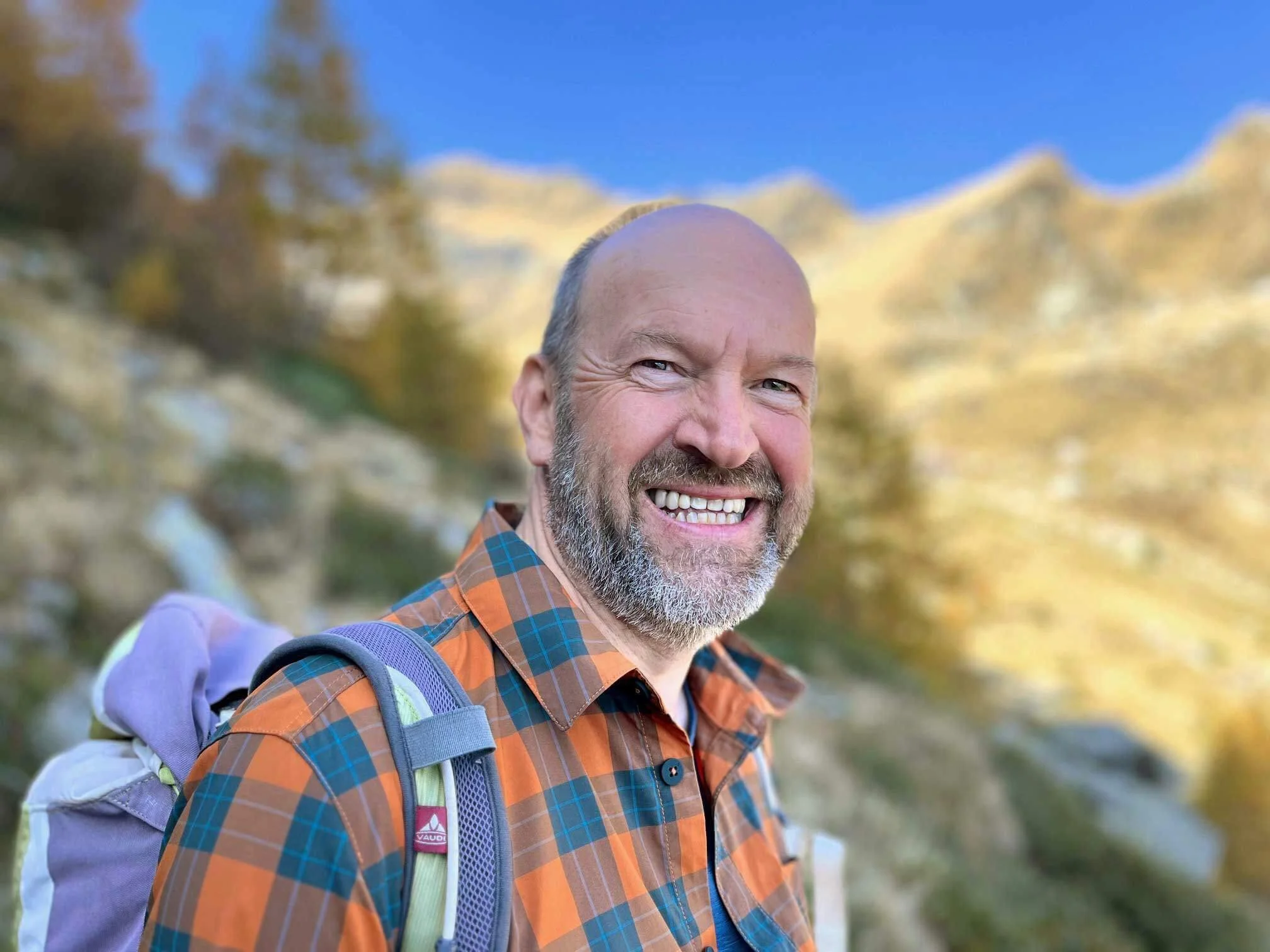 Ein lächelnder, älterer Mann mit grau-weißem Bart trägt einen Rucksack und steht outdoors mit einem bewölkten Himmel im Hintergrund.
