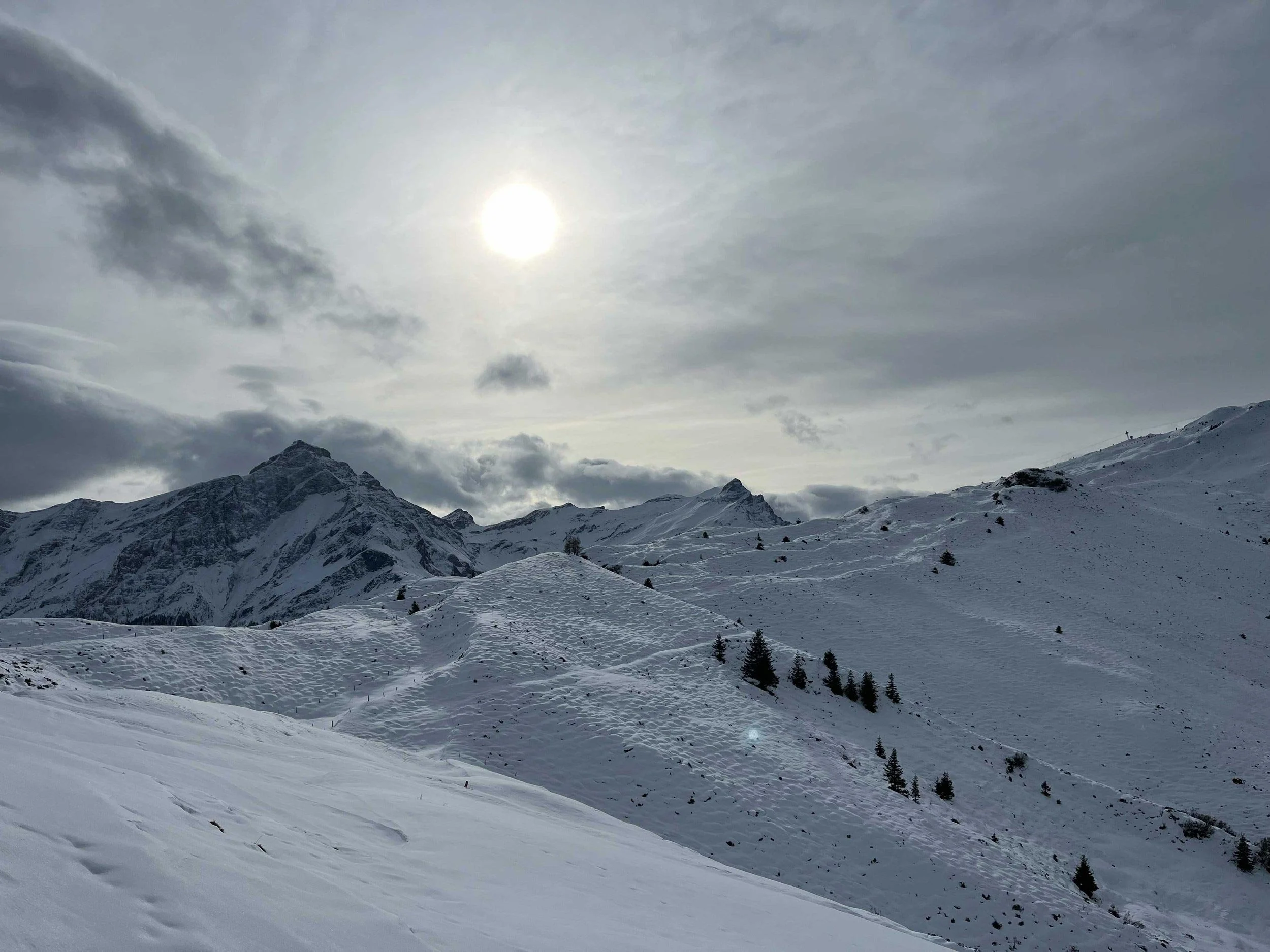 Berglandschaft mit schneebedeckten Gipfeln unter bewölktem Himmel, Sonne, die durch Wolken schiebt.