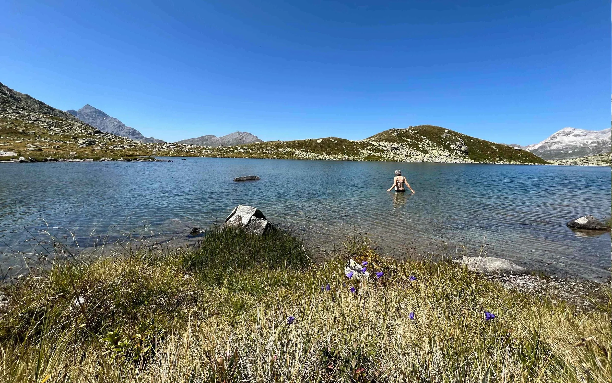 Eine Person steht im klaren Wasser eines Sees vor einer bergigen Landschaft mit grün bewachsenen Hügeln und schneebedeckten Gipfeln im Hintergrund unter einem blauen Himmel.