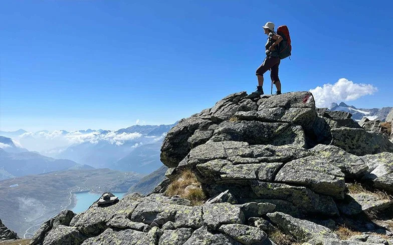 Ein Wanderer mit Rucksack auf einem Felsen in den Bergen mit Blick auf Wolken, Berge und einen See im Hintergrund.