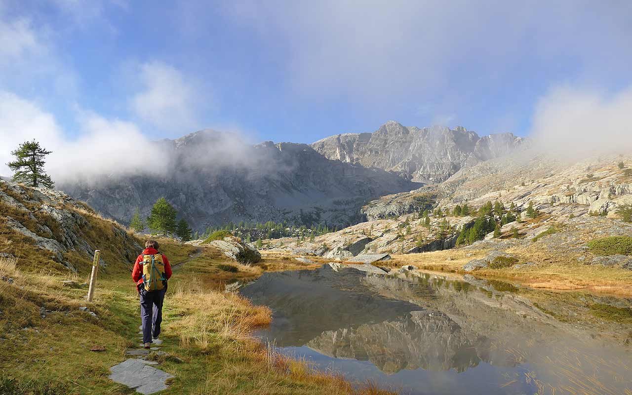 Ein Wanderer mit Rucksack geht an einem See in einer Bergregion vorbei, umgeben von Felsen und Bäumen, mit hoch aufragenden Bergen im Hintergrund und teils bewölktem Himmel.