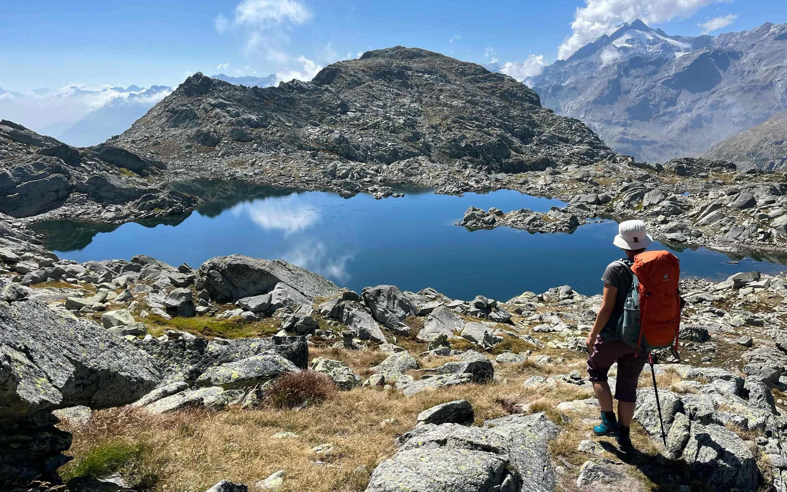Ein Wanderer mit Rucksack und Hut steht auf felsigem Gelände vor einem kleinen Bergsee in einer Berglandschaft mit hohen Gipfeln im Hintergrund.