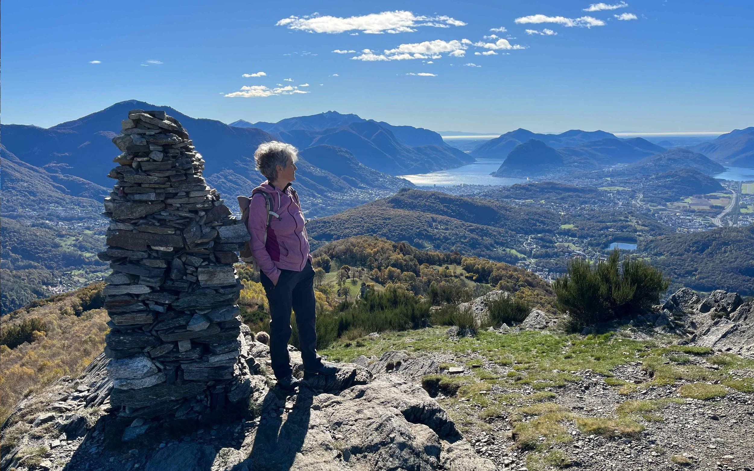 Eine ältere Frau mit grauem Haar, in pinker Jacke und dunklen Hosen, steht auf einem Berggipfel mit Blick auf eine Seen- und Berglandschaft bei schönem Wetter.