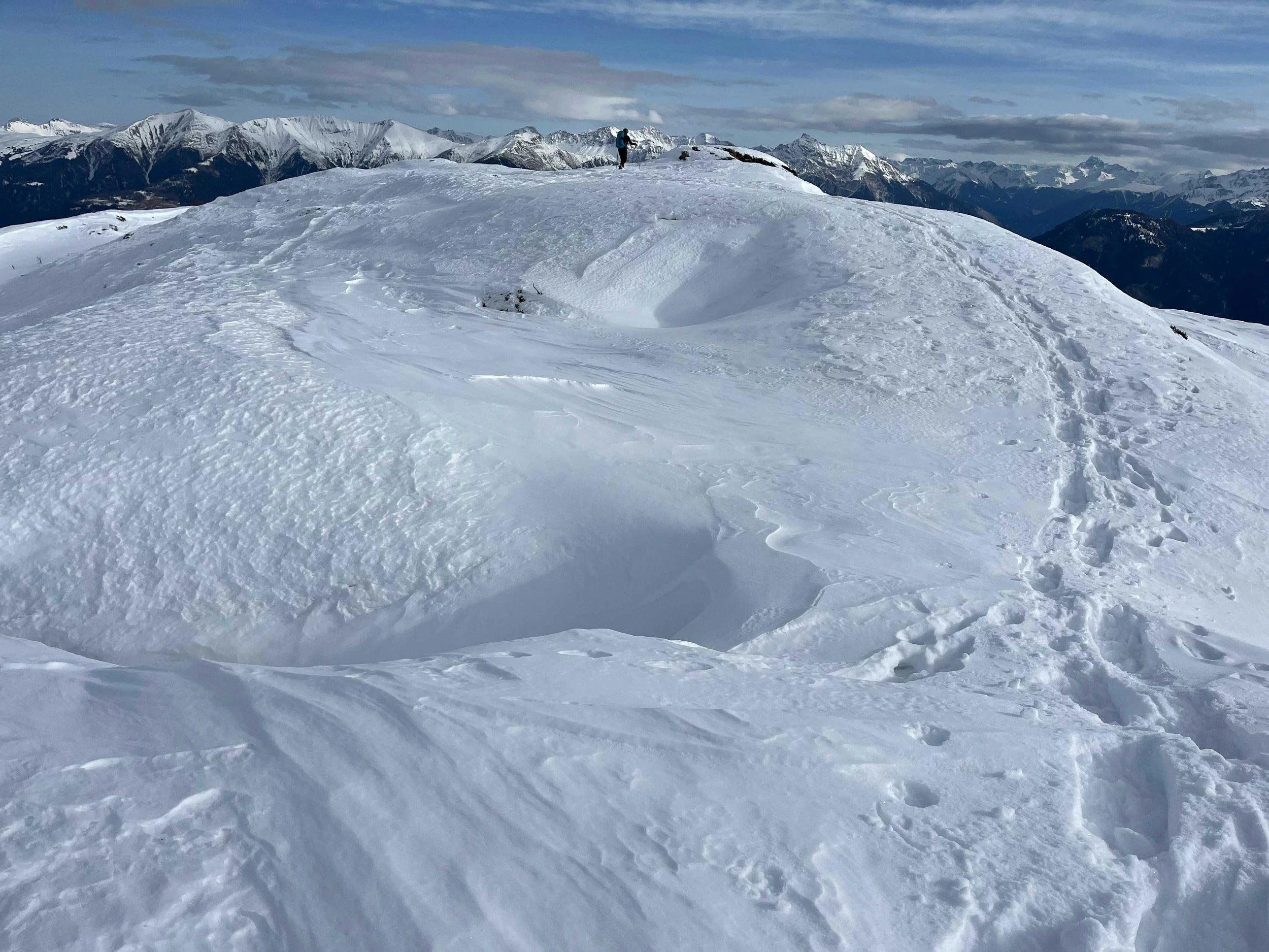 Ein Schneefeld in den Bergen mit einem Wanderer im Hintergrund, umgeben von schneebedeckten Gipfeln und Wolken am Himmel.