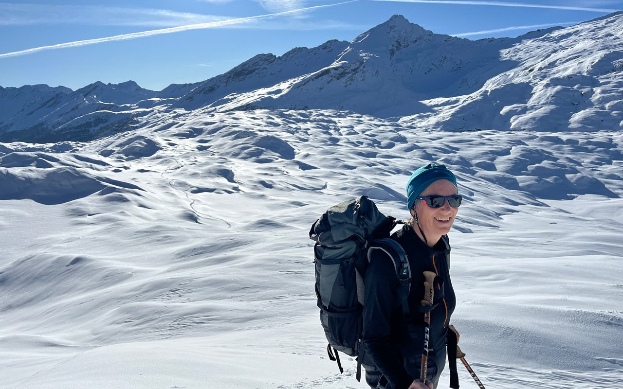 Eine lachende Frau mit Sonnenbrille, Mütze und Rucksack beim Schneeschuhwandern in einer verschneiten Berglandschaft mit hohen Gipfeln unter blauem Himmel.