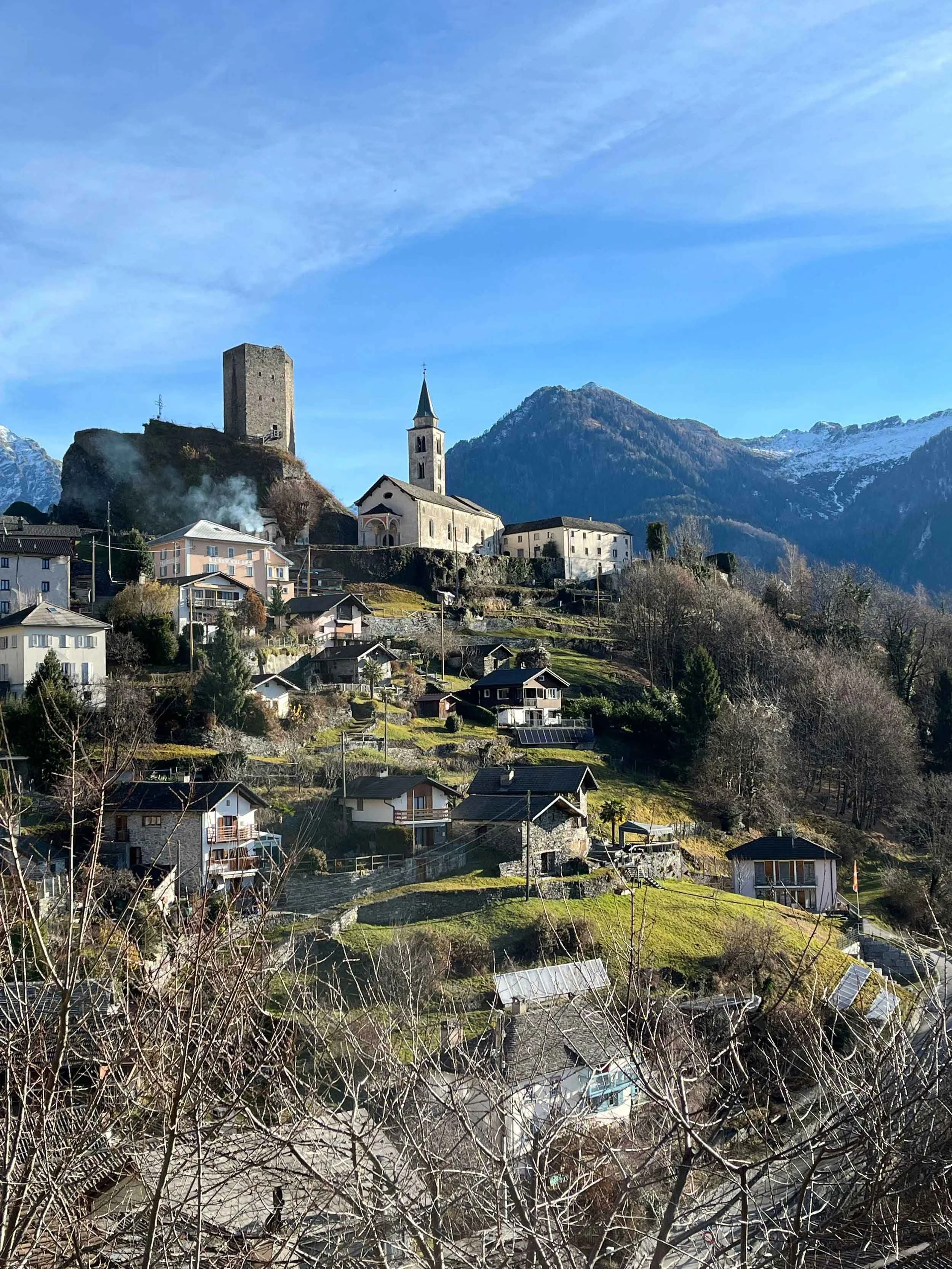 Bergdorf mit Kirche, Burgturm und schneebedeckten Gipfeln im Hintergrund, sonniger Tag.