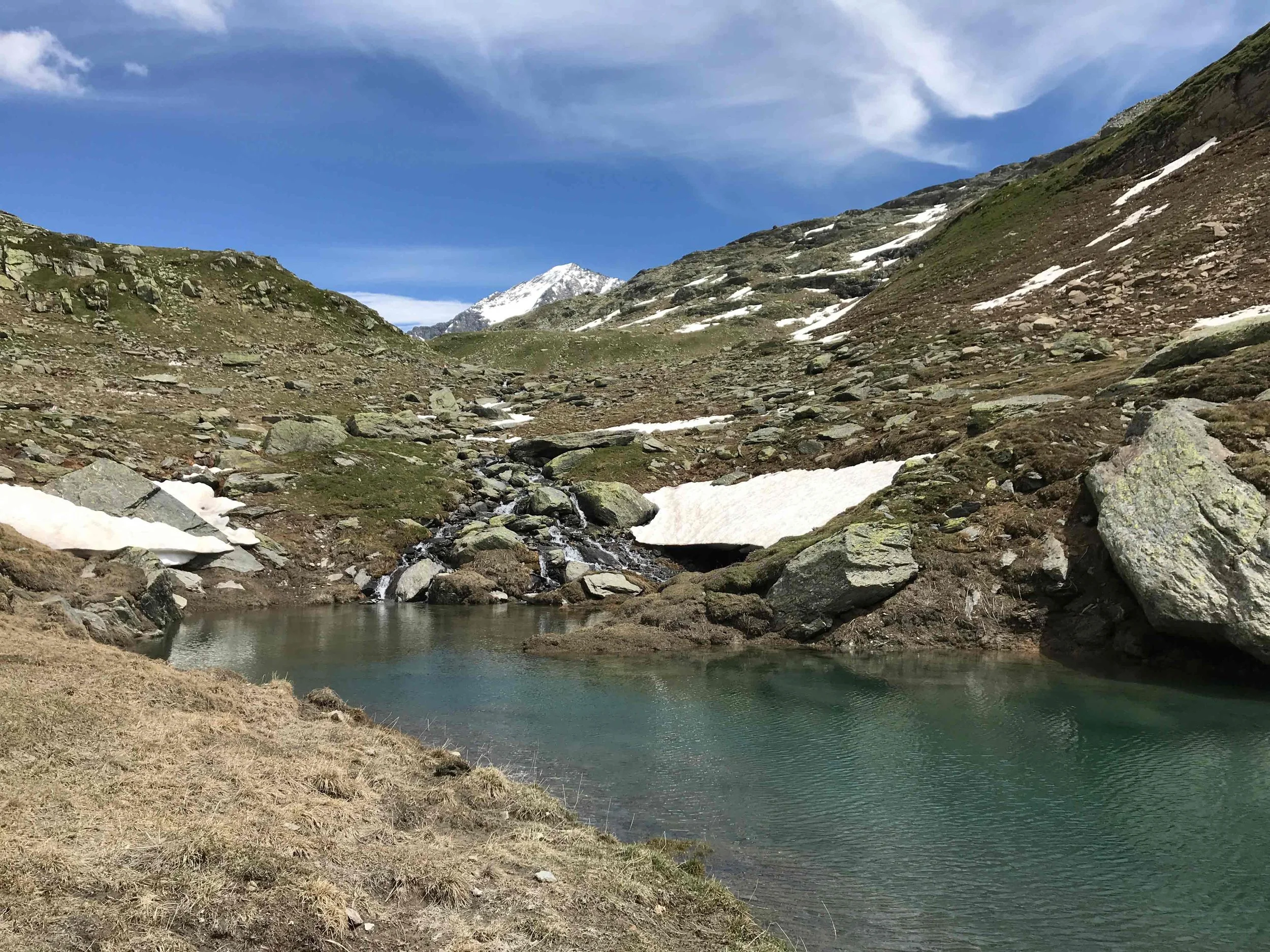 Bergsee in den Bergen mit Wasser, Felsen, Schneeresten und einem hohen Gipfel im Hintergrund, unter einem blauen Himmel mit Wolken.