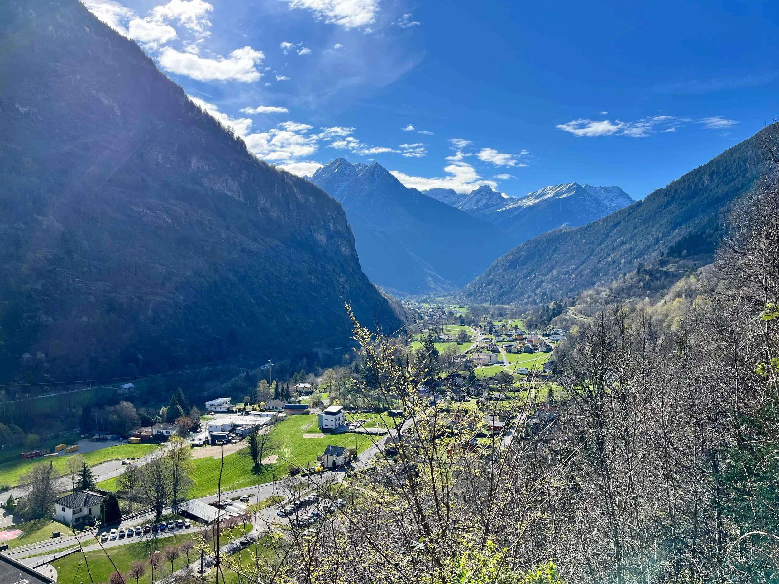 Bergdorf in einer Talansicht mit grünen Feldern, waldigen Hügeln und schneebedeckten Bergen im Hintergrund, bei sonnigem Himmel mit wenigen Wolken.