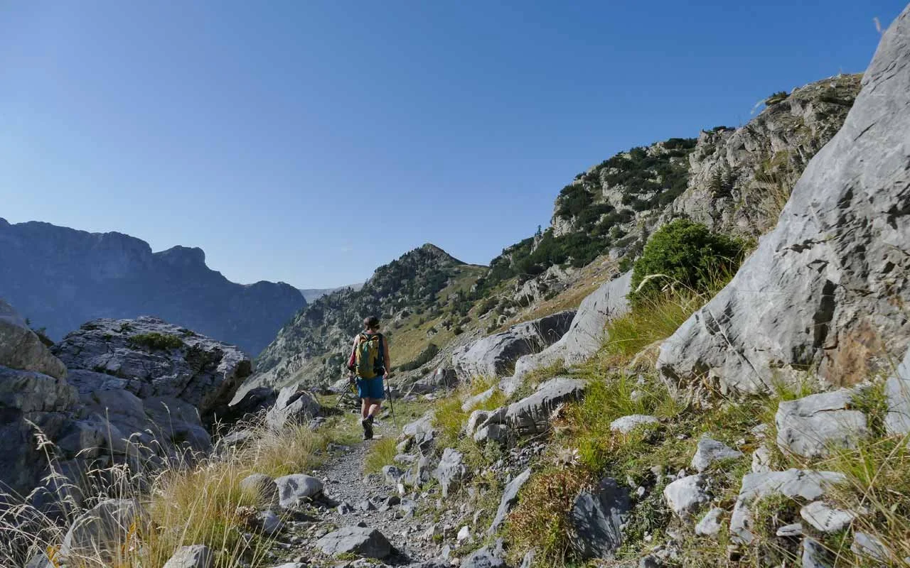 Ein Wanderer mit Rucksack und Wanderstöcken wandert durch ein felsiges Gebirge unter klarem blauen Himmel.