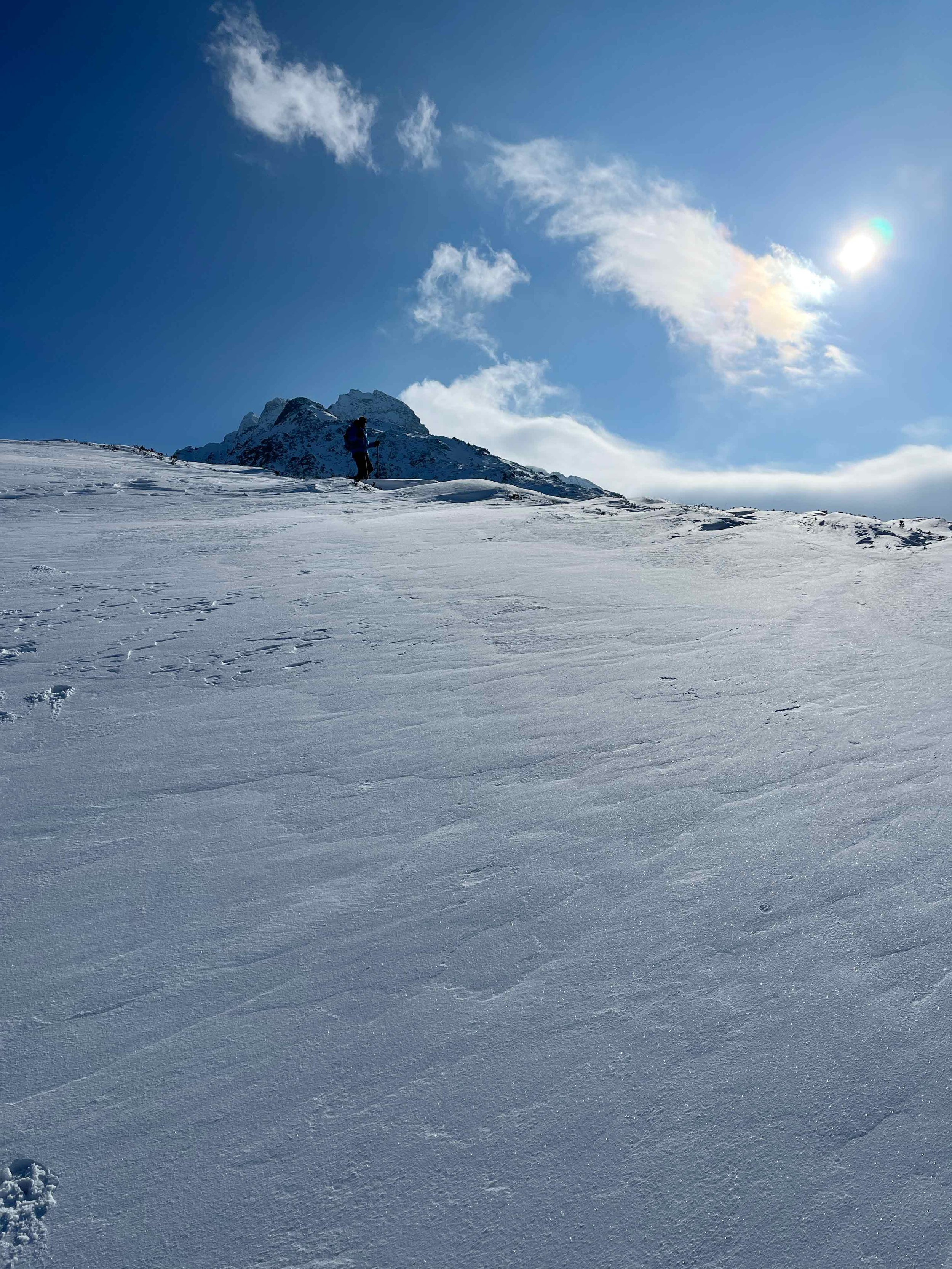 Ein Schneeschuhwanderer steht auf schneebedeñter Oberfläche mit Berg im Hintergrund und sonnigem Himmel.