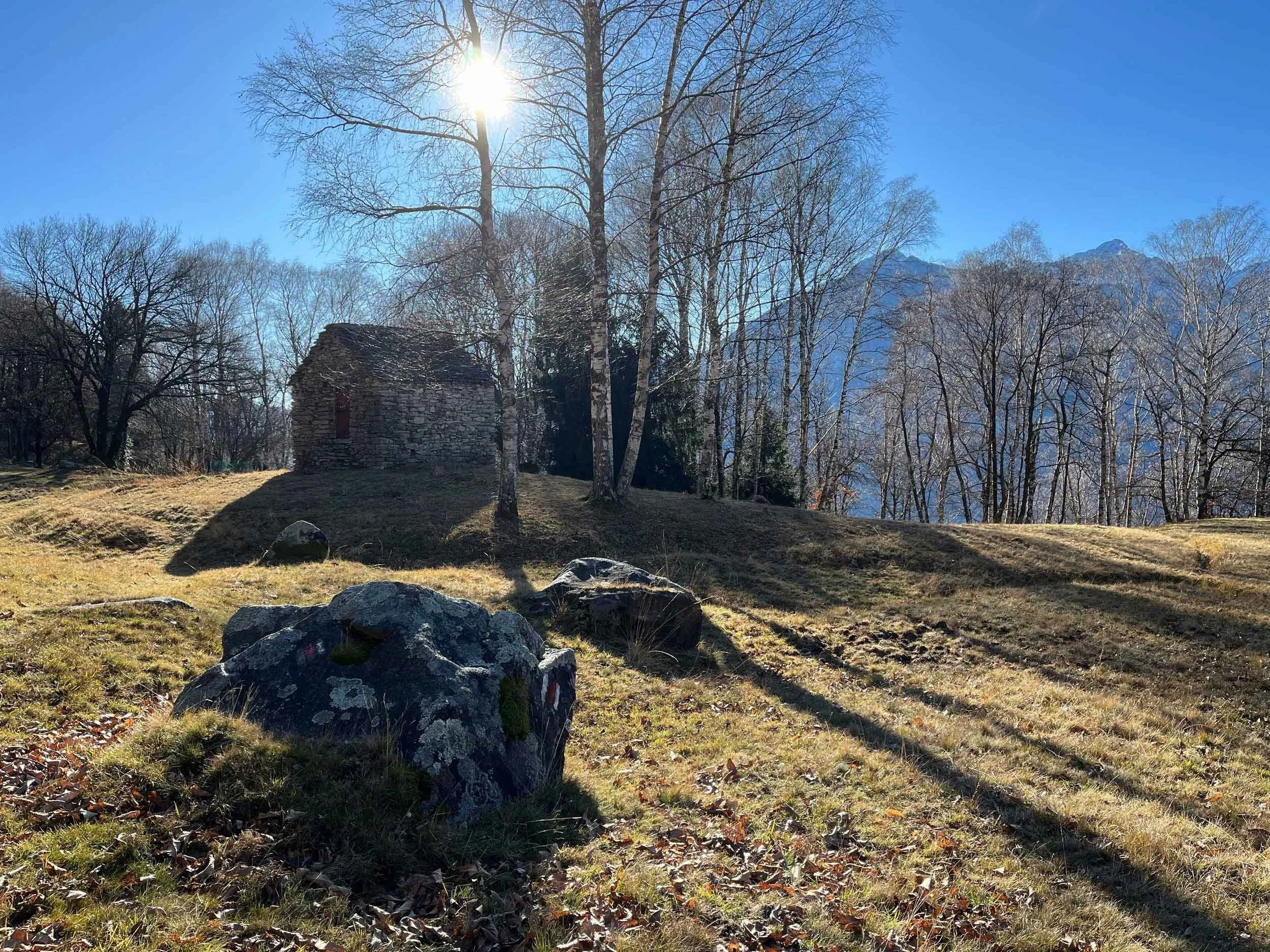Ein sonniger Wintertag in den Bergen mit kahlen Bäumen, einer kleinen Stein- hütte im Vordergrund und schneebedeckten Gipfeln im Hintergrund.