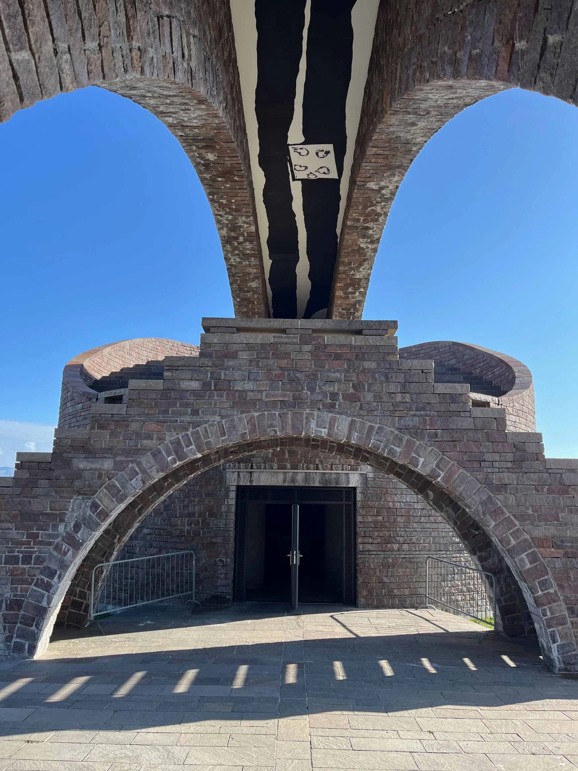 Blick auf einen Eingang mit großen Backsteinbögen und einer Doppeltür, im Vordergrund ein gepflasterter Vorplatz mit Schatten. Das Gebäude hat eine alte Architektur mit Zinnen und einem simulierten Bogengang.