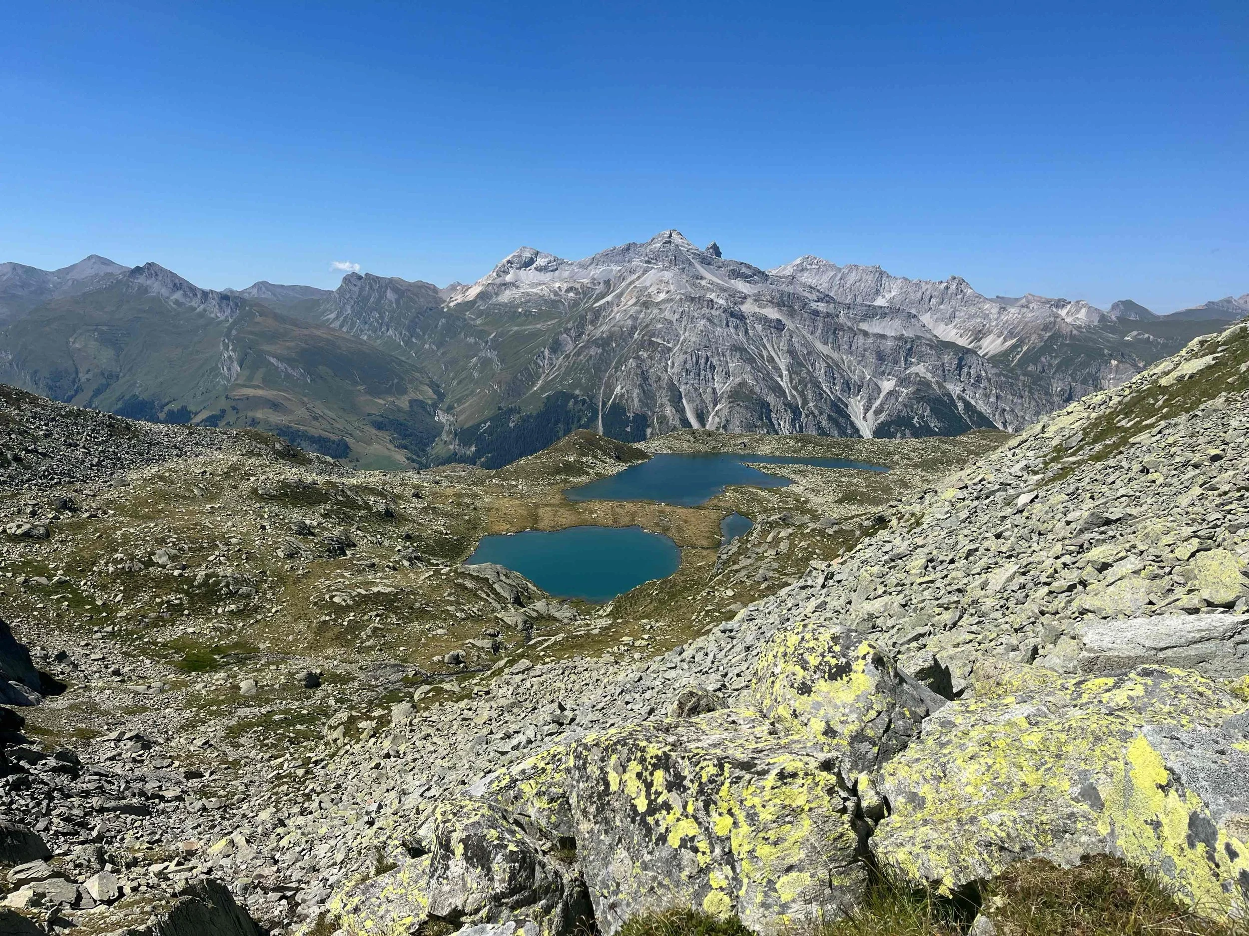 Bergsee inmitten von Felsen und Bergen mit schneebedeckten Gipfeln im Hintergrund bei blauem Himmel.