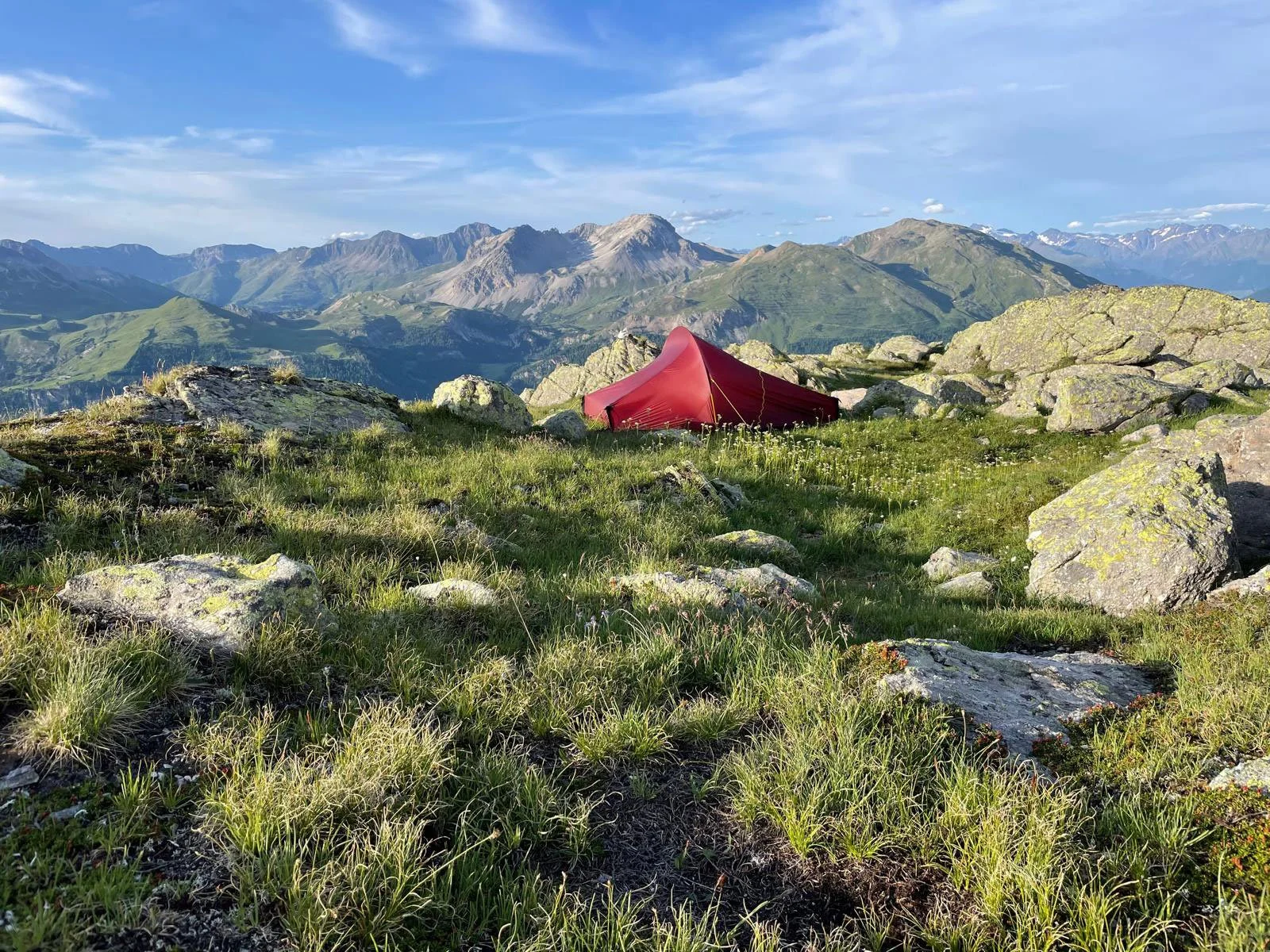 Ein rotes Zelt liegt in einer grünen, rocky Berglandschaft mit hohen Bergen und Wolken im Himmel im Hintergrund.