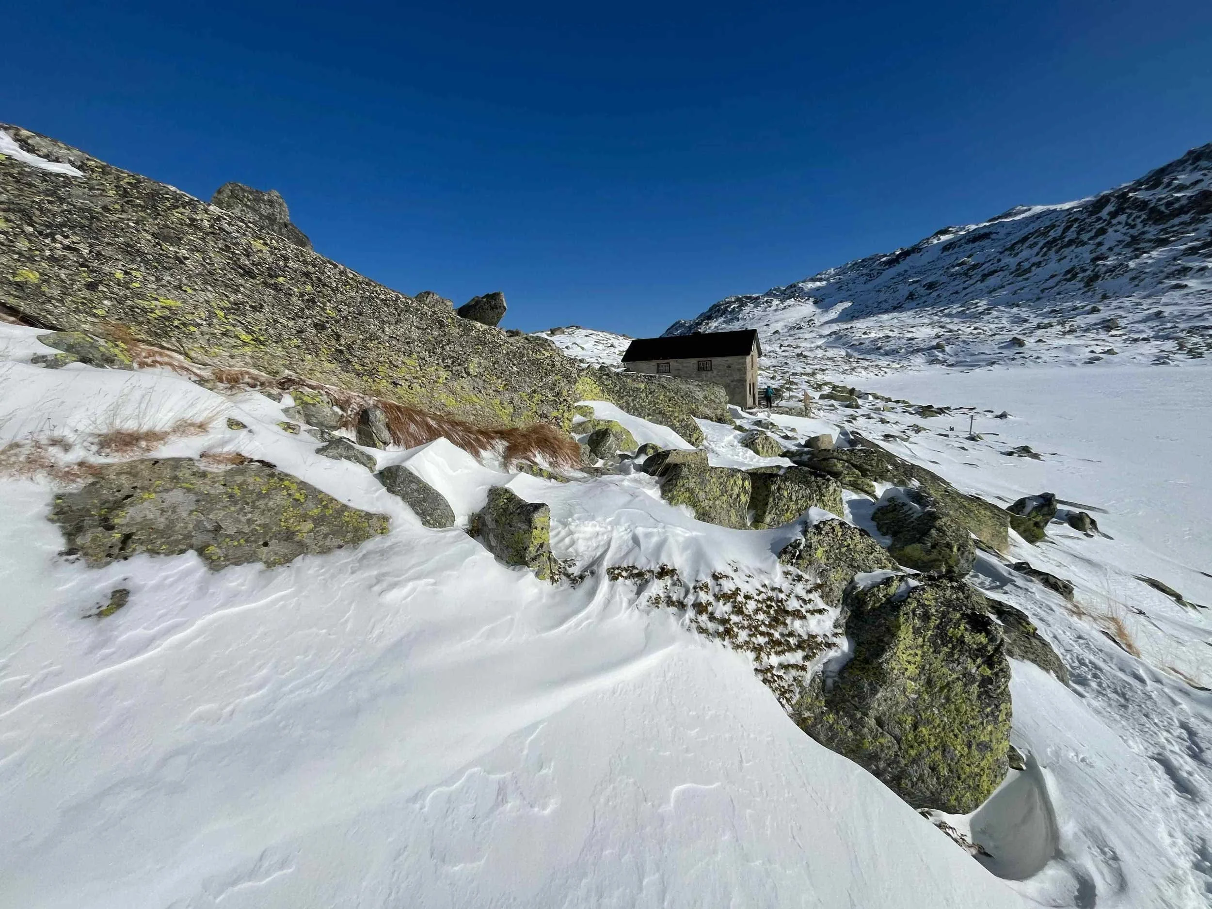 Ein verschneiter Bergsee mit einer einsamen Hütte im Hintergrund, umgeben von schneebedeckten Bergen und einem klaren blauen Himmel.