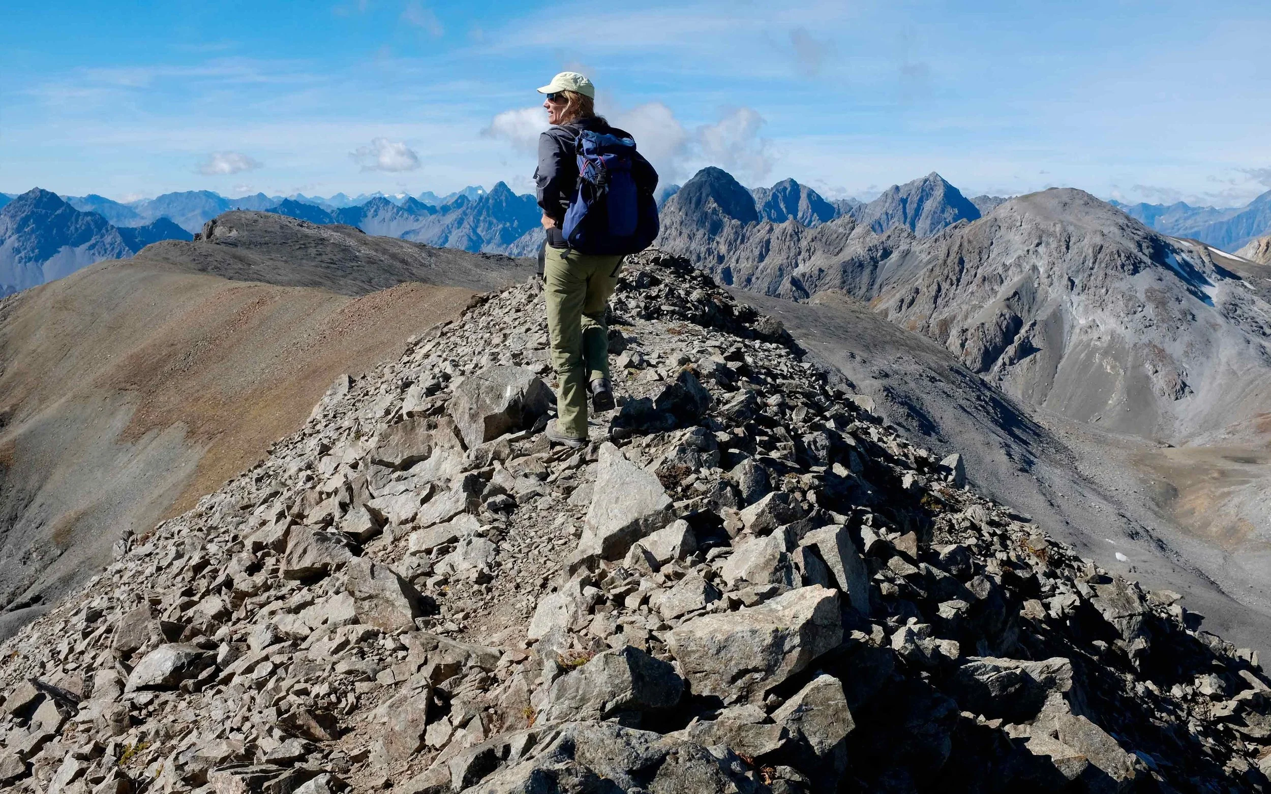 Eine Person wandert auf einem felsigen Bergkamm, umgeben von hohen Bergen unter einem blauen Himmel mit einigen Wolken.