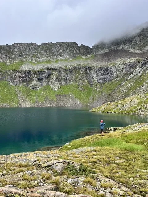 Ein einzelner Mensch fischt an einem klaren Bergsee, umgeben von grünen Hügeln und steilen Felswänden bei bewölktem Himmel.