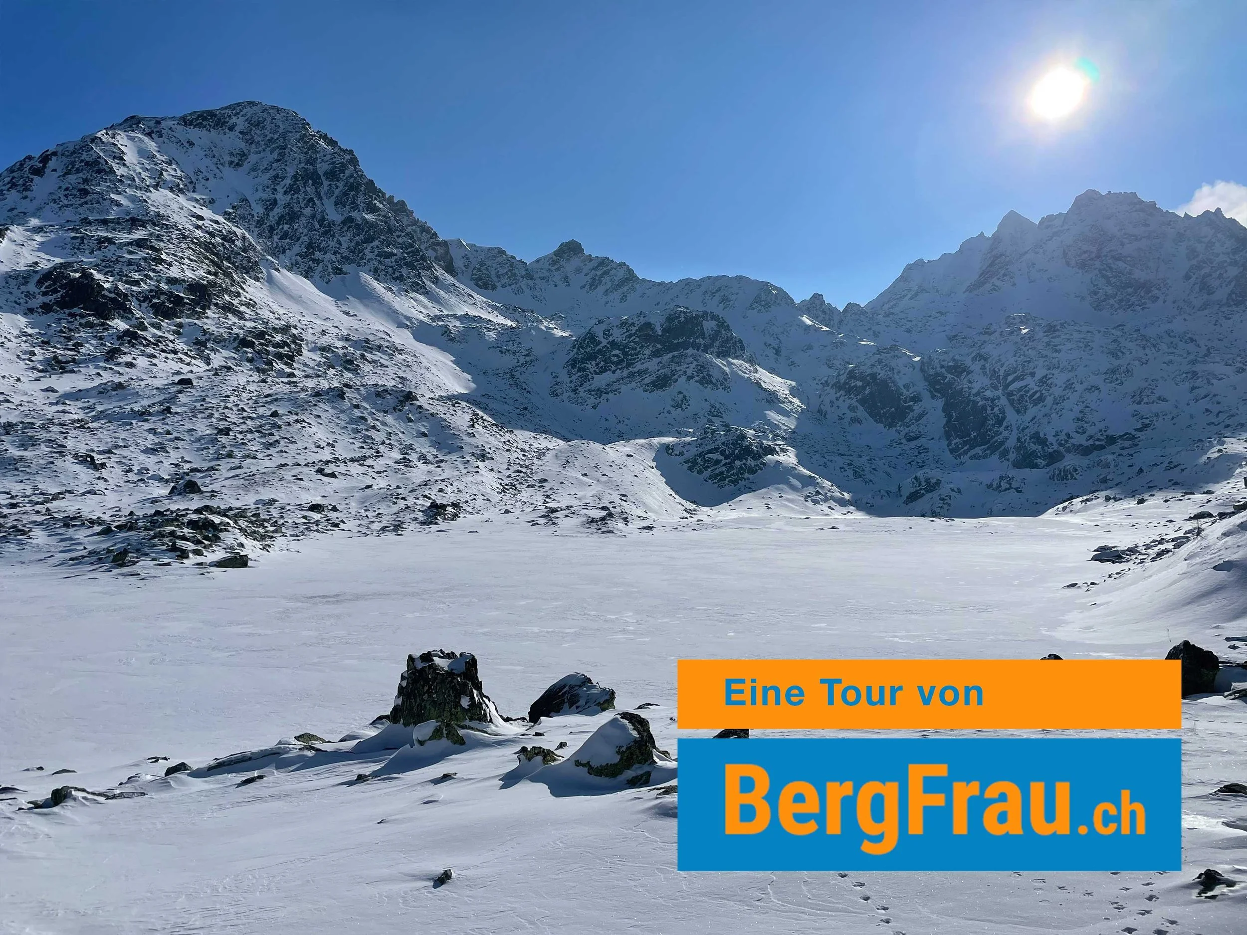 Eine verschneite Berglandschaft mit hohen, schneebedeckten Gipfeln, einer gefrorenen See und einem klaren blauen Himmel mit der Sonne, im Vordergrund Steine im Schnee. Text auf dem Bild: 'Eine Tour von BergFrau.ch'.