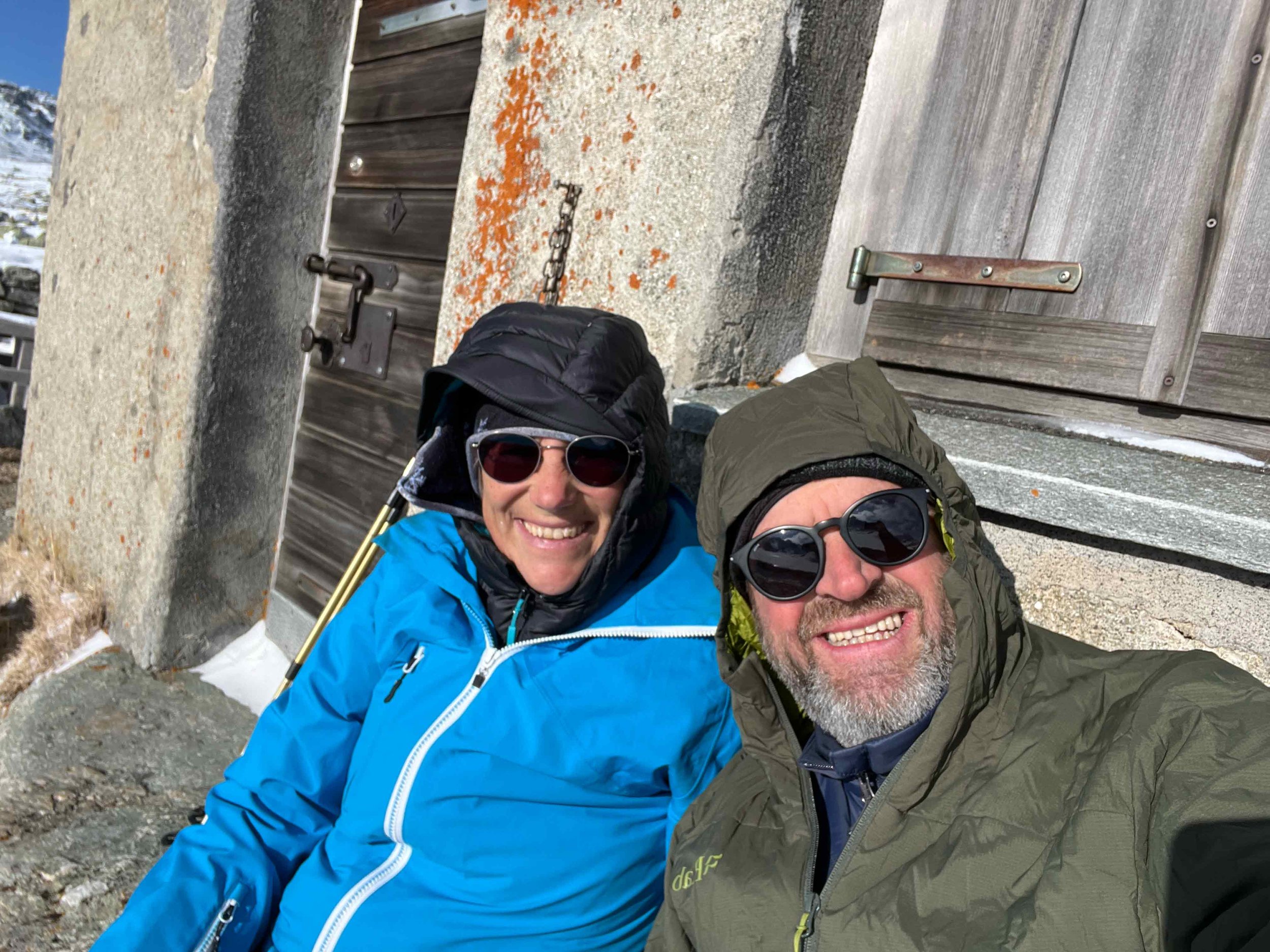 Ein Mann und eine Frau tragen Outdoor-Bekleidung und Sonnenbrillen, sie machen ein Selfie vor einem alten Holz- und Steinschuppen in einer Bergregion mit Schnee im Hintergrund.