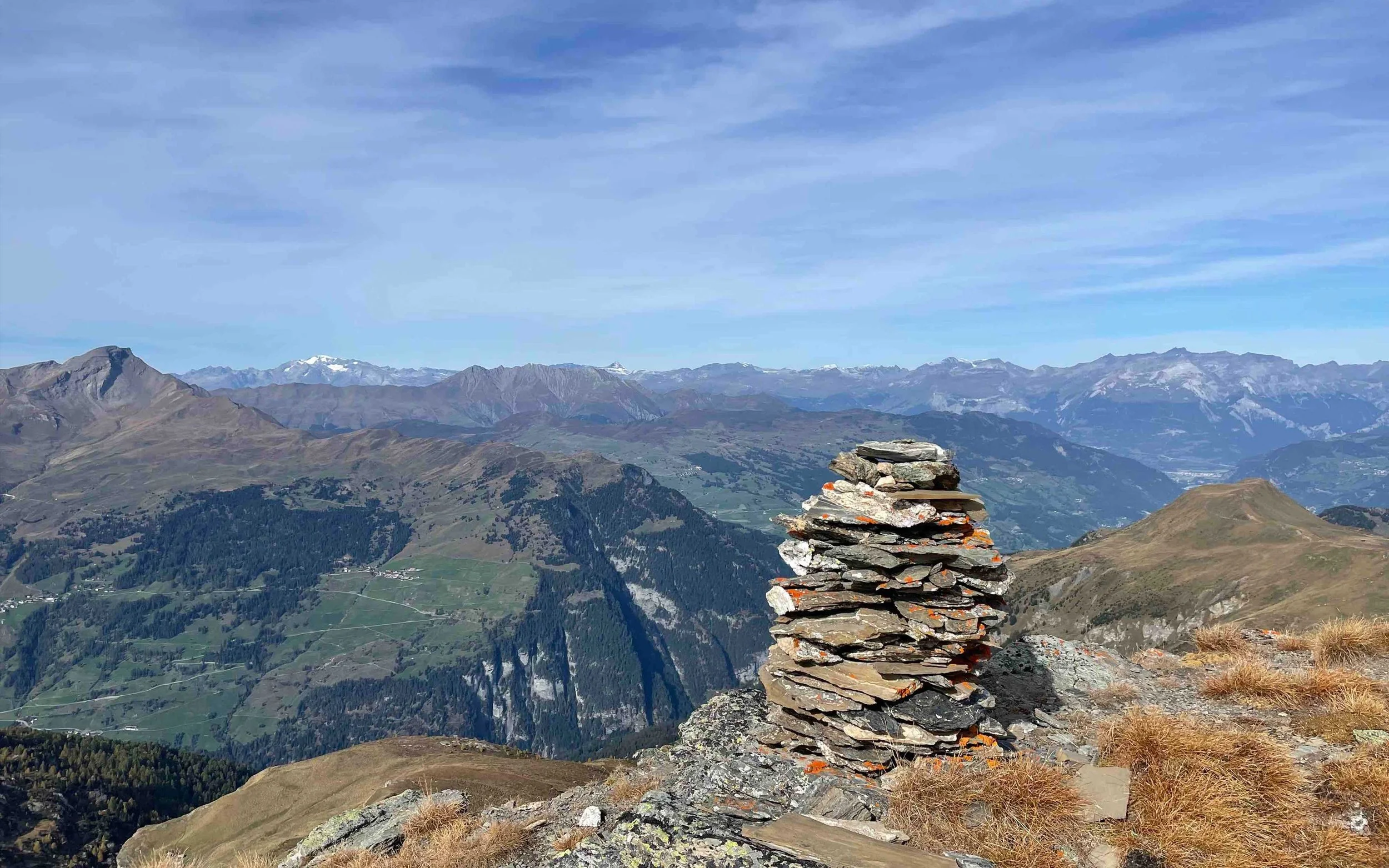 Bergpanorama mit steinernem Gipfelkreuz auf einem Gipfel, im Hintergrund schneebedeckte Berge unter blauem Himmel.