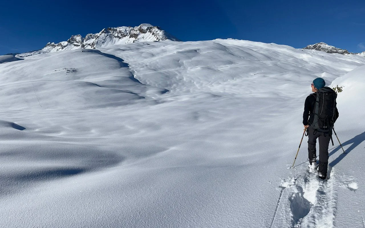 Ein Winterwanderer mit Rucksack und Wanderstöcken in schneebedechter Berglandschaft, blauer Himmel im Hintergrund.