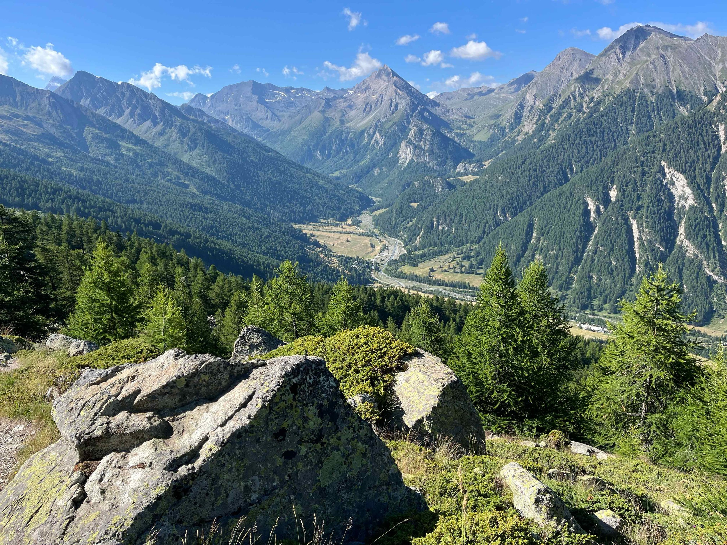 Berglandschaft mit grünen Wäldern, hohen Bergen und einem Fluss im Tal unter einem blauen Himmel mit Wolken.