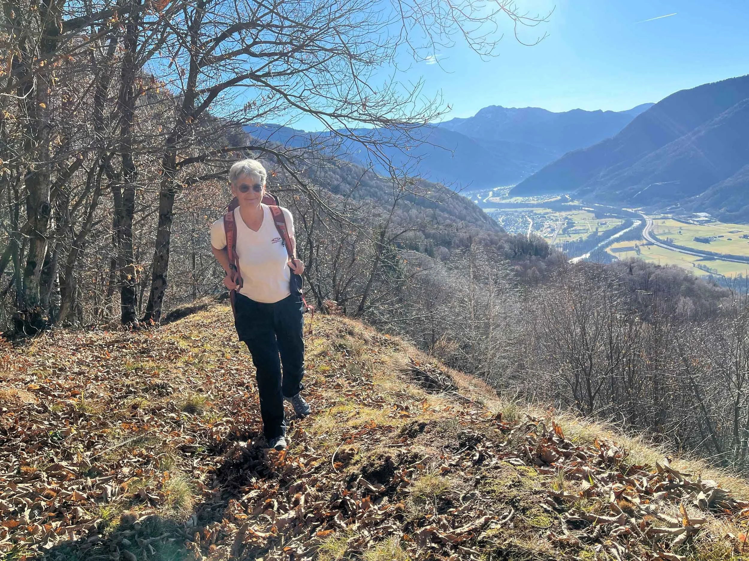 Eine ältere Frau mit Sonnenbrille und Rucksack wandert auf einem Pfad durch den Wald, im Hintergrund sind Berge und ein Fluss sichtbar.