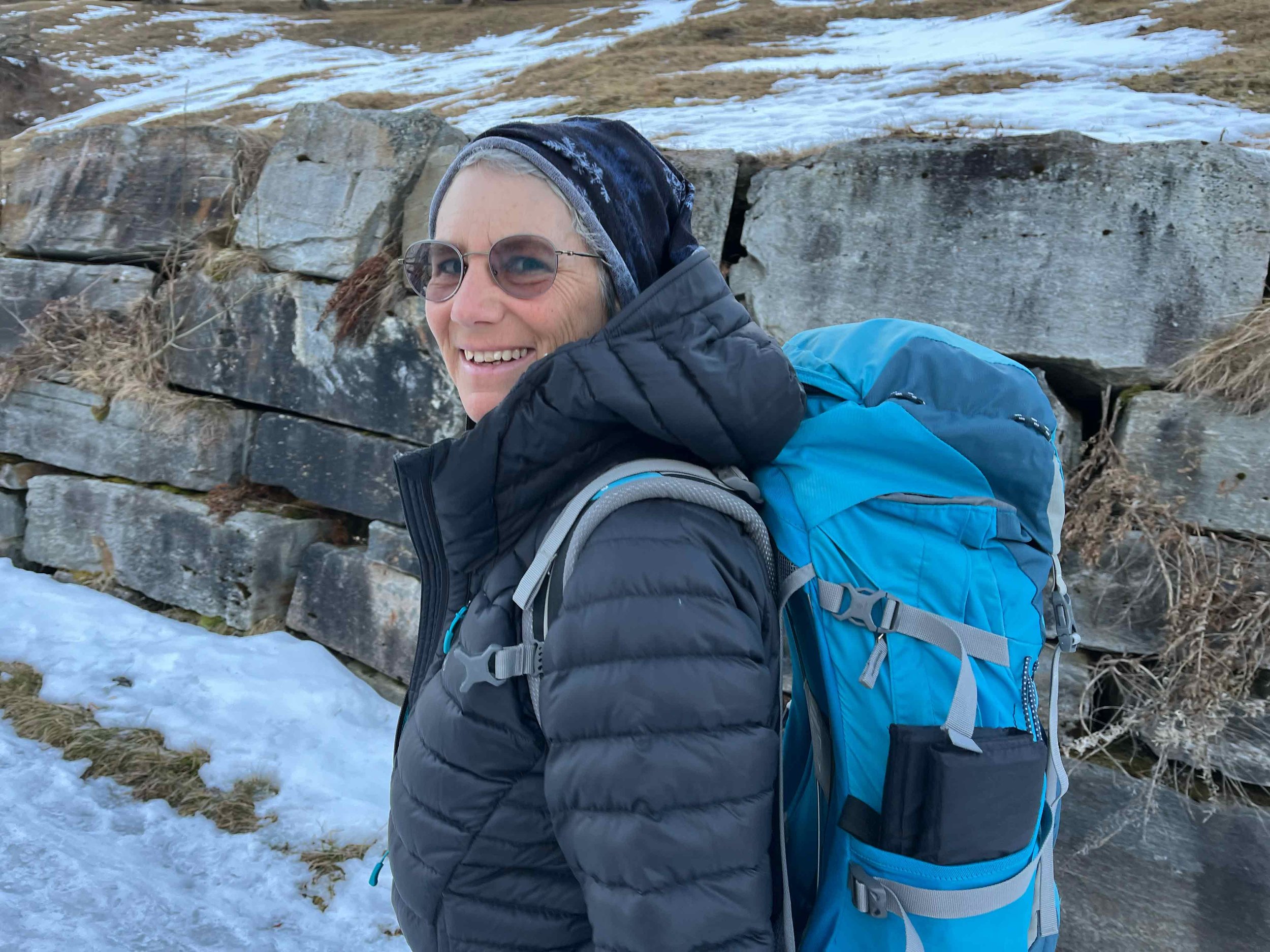 Eine Frau mit Sonnenbrille, schwarzer Jacke und blauer Rucksack beim Wandern in verschneiter Natur.