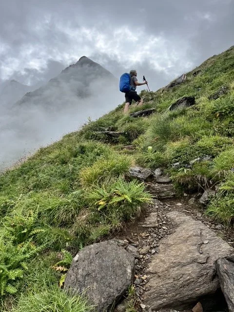 Person mit Wanderrucksack auf steilem, nassen Bergpfad, im Hintergrund Wolken und Nebel, Gipfel im Hintergrund