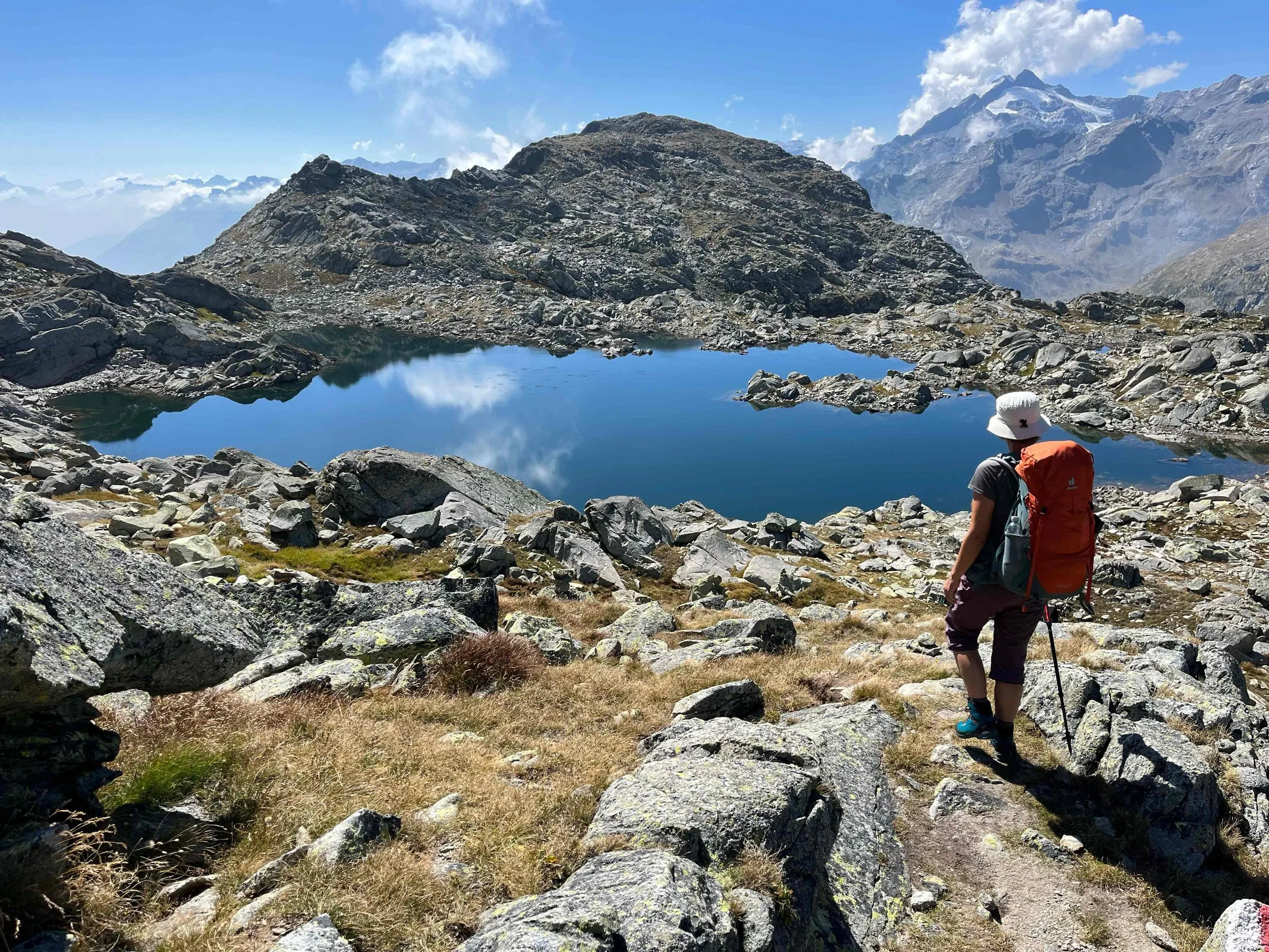 Ein Wanderer mit Rucksack und Hut auf einem felsigen Gebirgsweg, Blick auf einen ruhigen Bergsee und schneebedeckte Gipfel im Hintergrund, bei sonnigem Himmel.