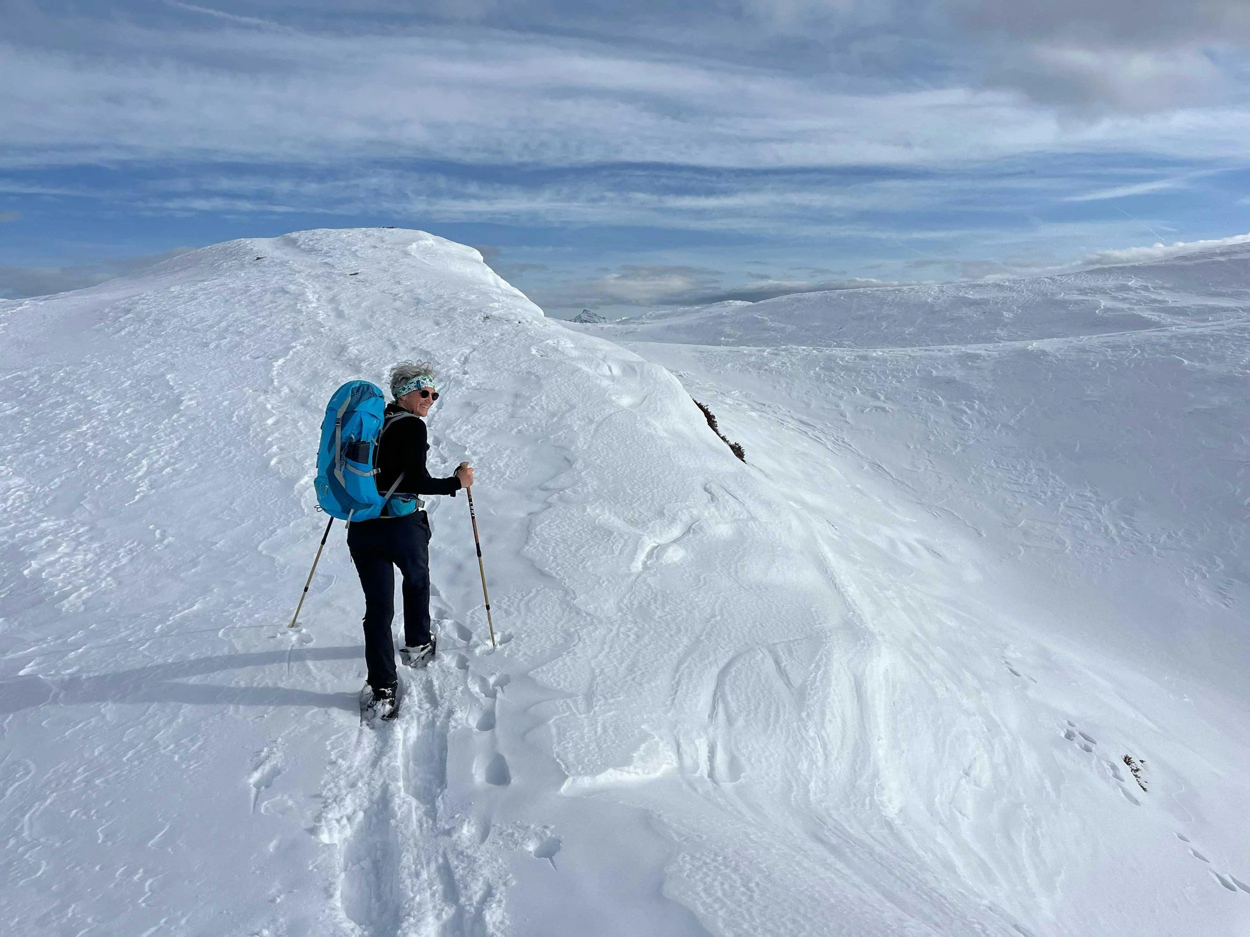 Eine Person mit Sonnenbrille, Winterbekleidung und Rucksack wandert auf verschneitem Bergmassiv, umgeben von weißen Schneehügeln und einem blauen Himmel mit Wolken.