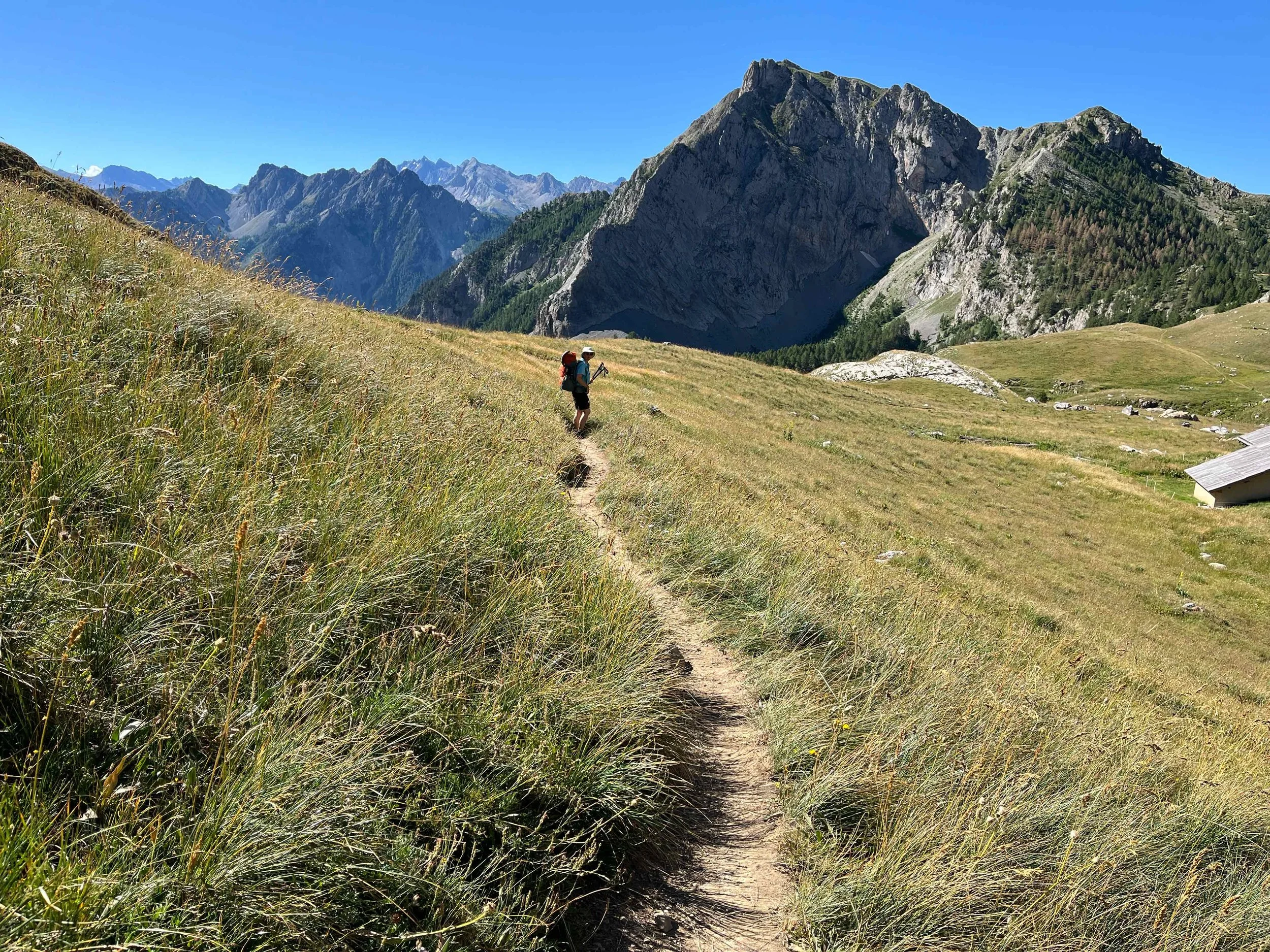 Zwei Wanderer auf einem schmalen Pfad in einer grünen bergigen Landschaft mit hohen Bergen im Hintergrund bei sonnigem Himmel.