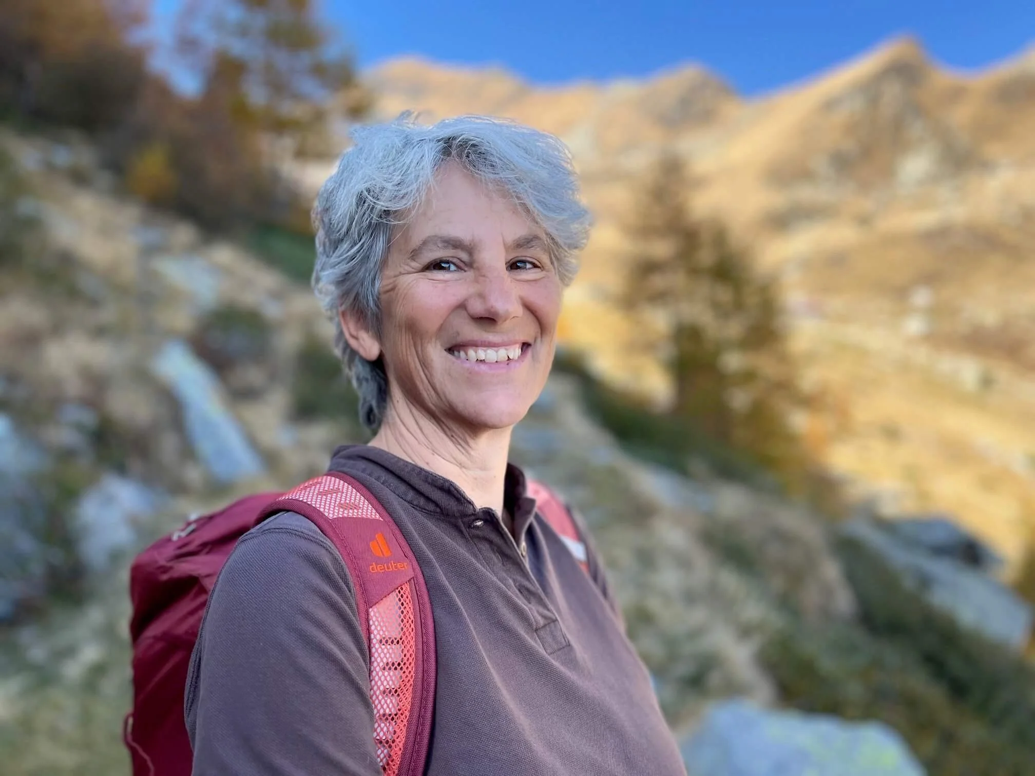 Eine lächelnde ältere Frau mit kurzen grauen Haaren, trägt eine braune Jacke und einen roten Rucksack, im Hintergrund ist eine Naturkulisse mit Bäumen und einem blauen Himmel.