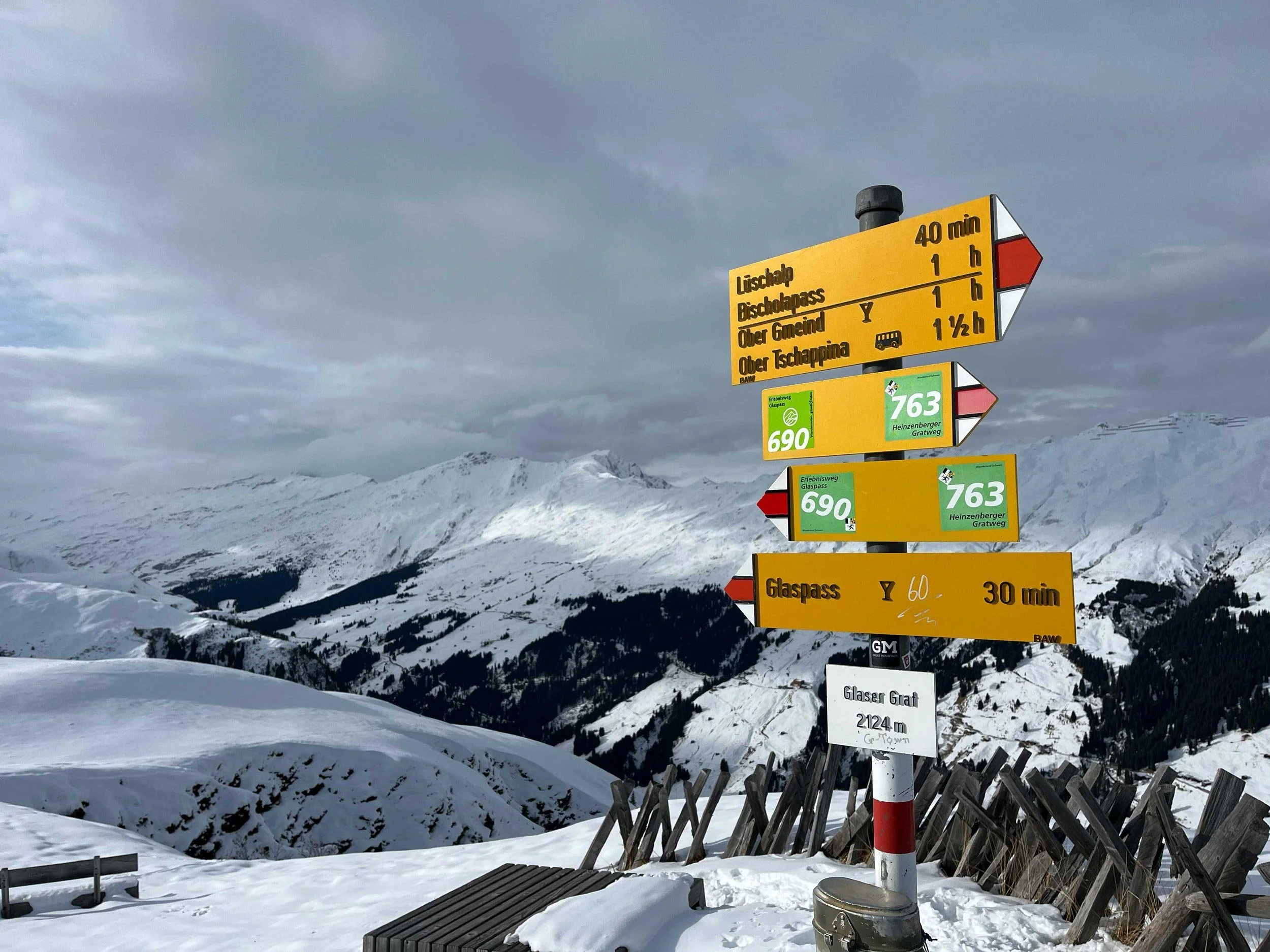 Schneebedeckte Berge und Skilifte mit Richtungs- und Zeitangaben in deutscher Sprache auf gelben und grünen Schildern, an einem Winterhimmel. 
