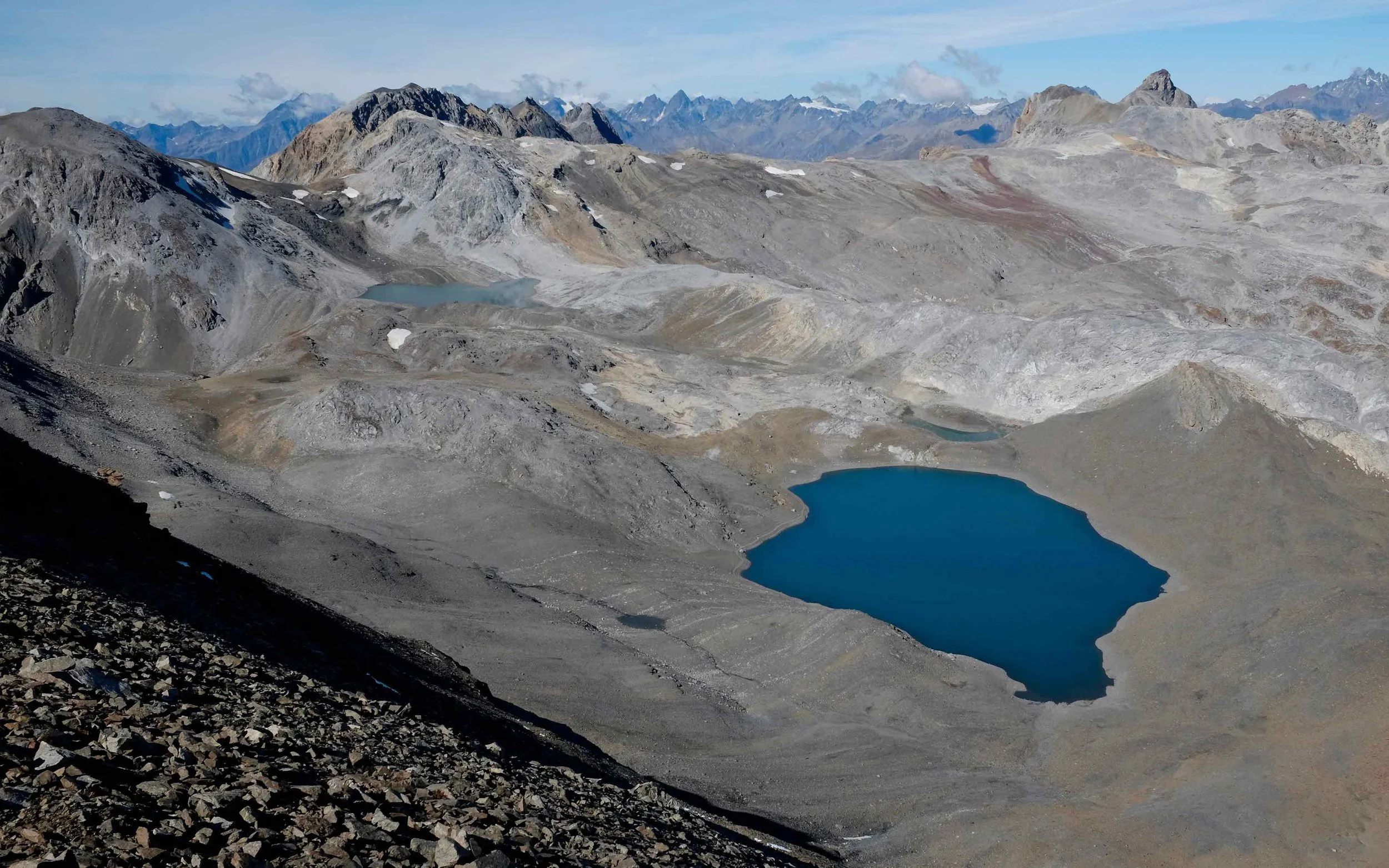 Bergsee in der alpinen Landschaft mit Gletscherresten, umgeben von grauen und braunen Bergen unter einem bewölkten Himmel.