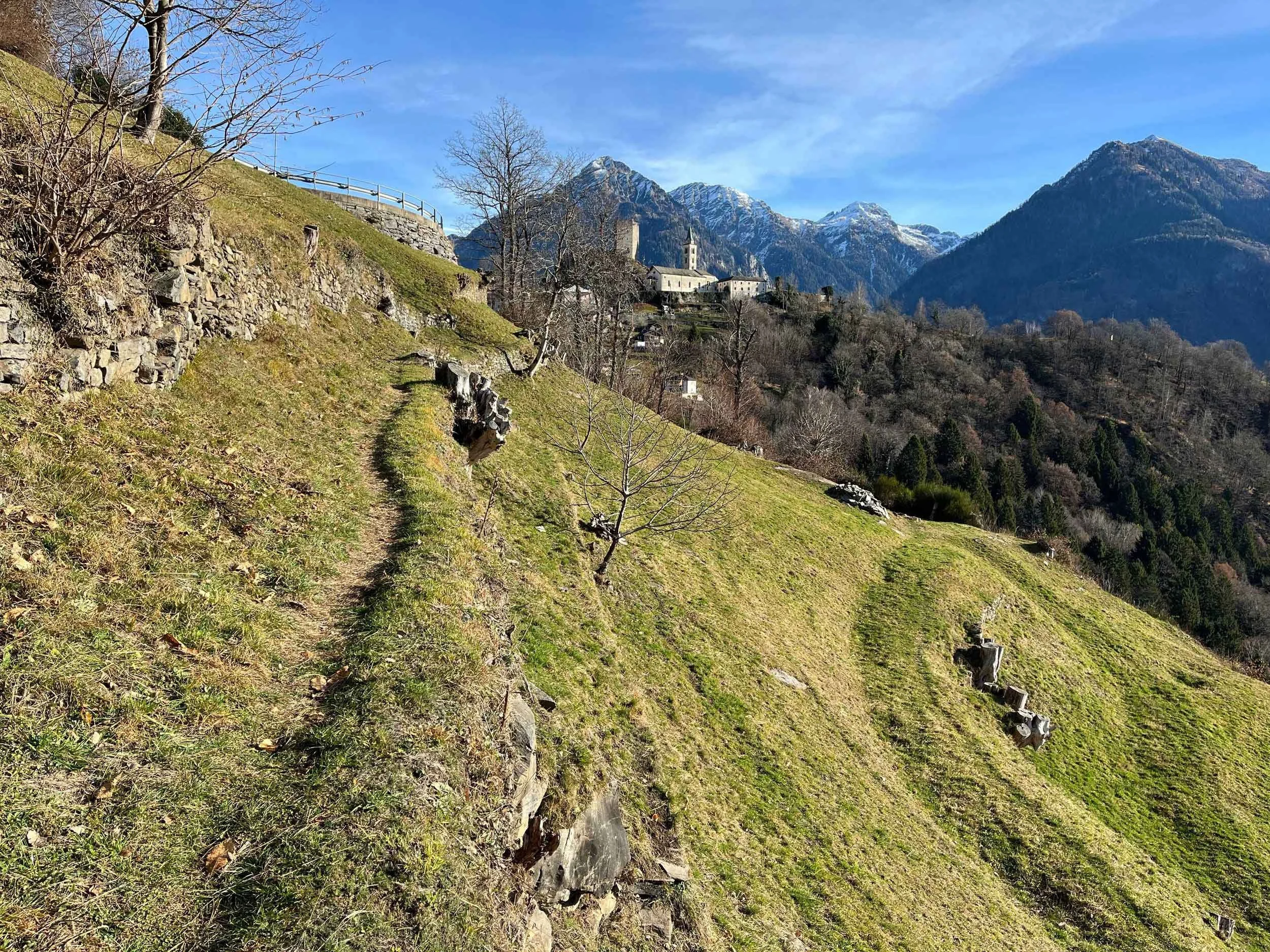 Bergdorf mit Kirche auf einem Hügel, umgeben von Bergen und Bäumen, sonniger Tag, klarer blauer Himmel.