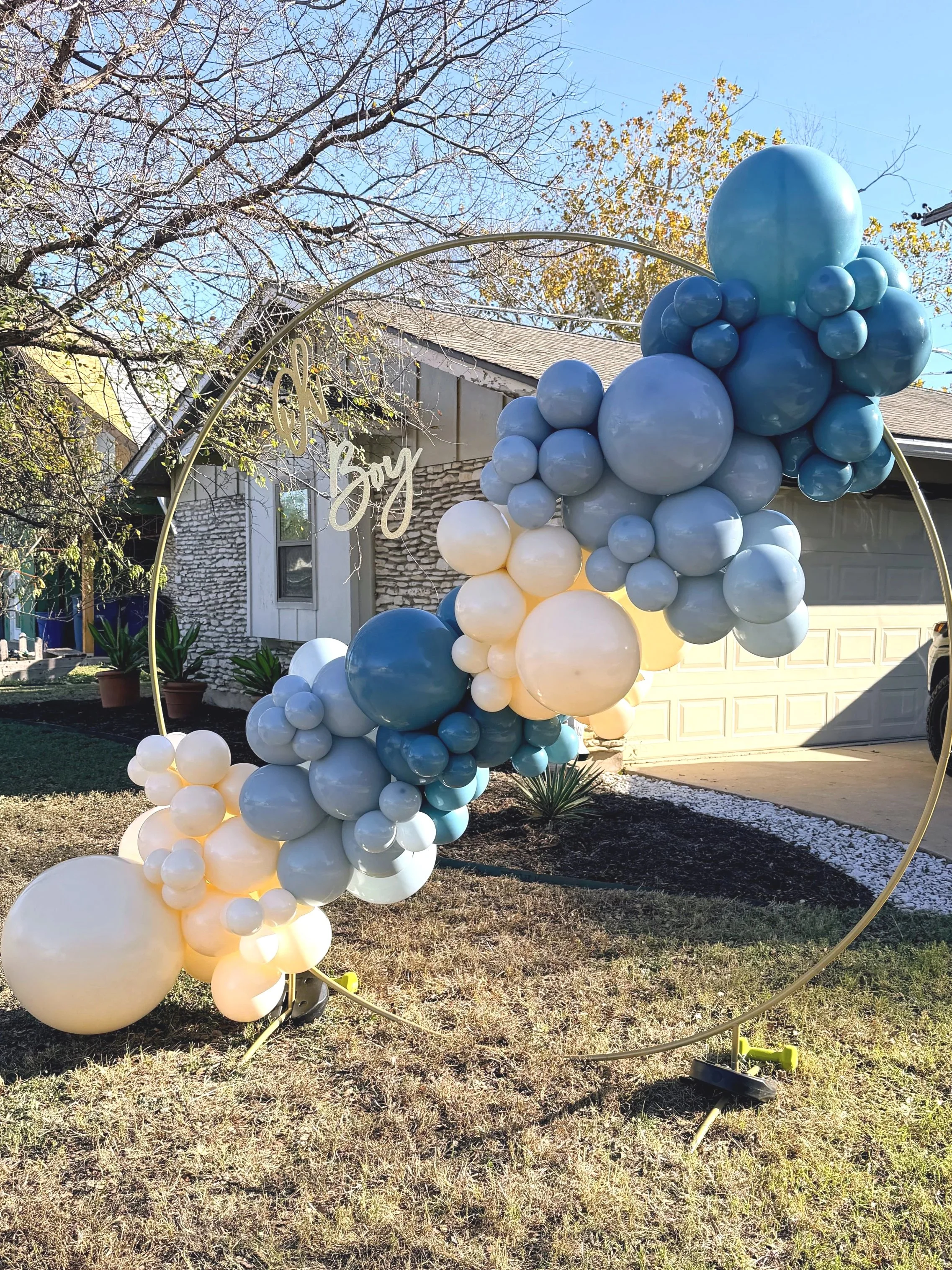 Custom blue and white balloon garland on a gold hoop backdrop for a baby boy event, installed outdoors by 512BalloonMom.