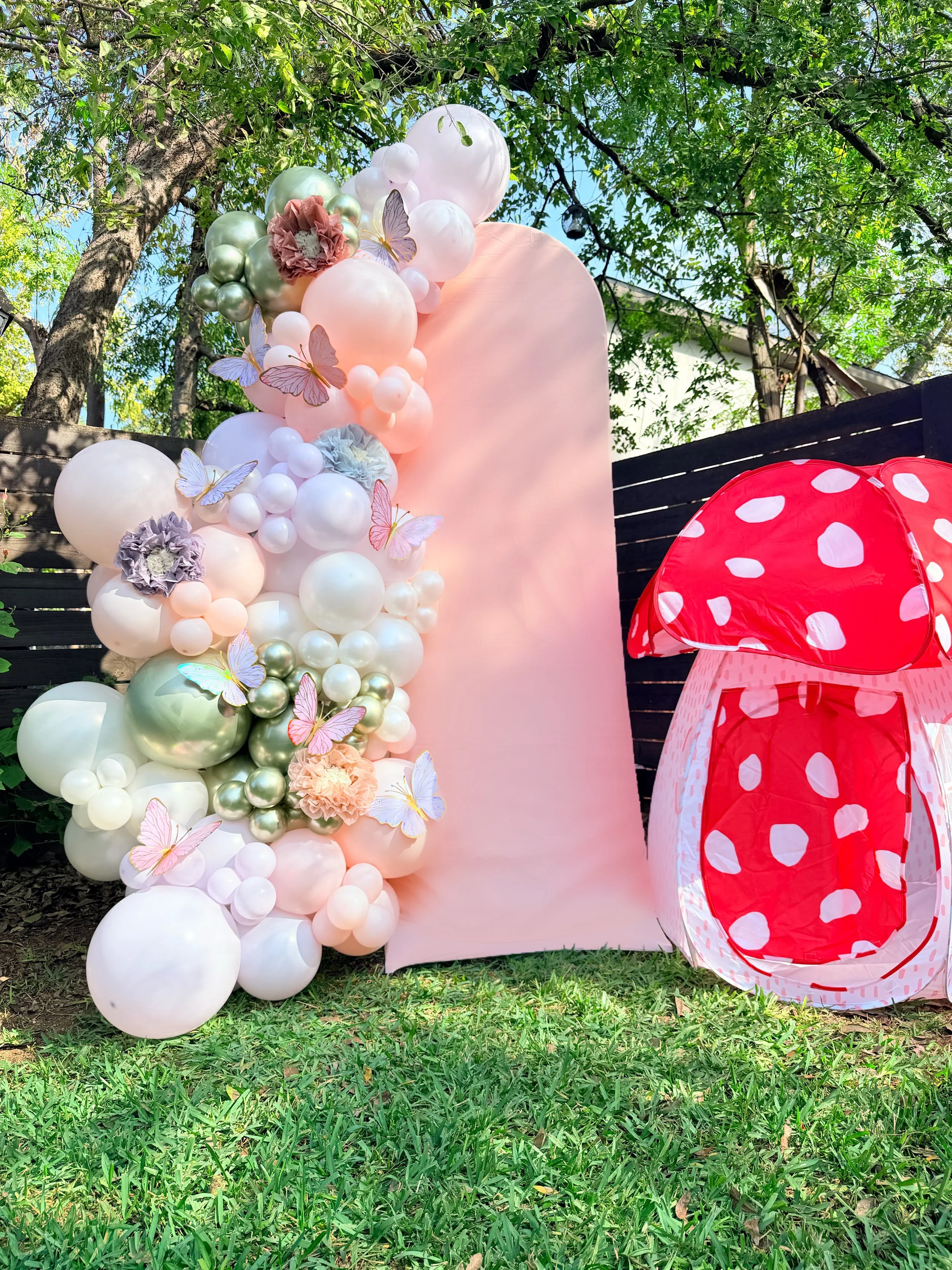 Colorful outdoor decoration with a large pink backdrop, white and pastel-colored balloons, paper flowers, and butterfly cutouts on the left side, and a red and white polka-dotted mushroom-shaped tent on the right, set on green grass with trees in the background.