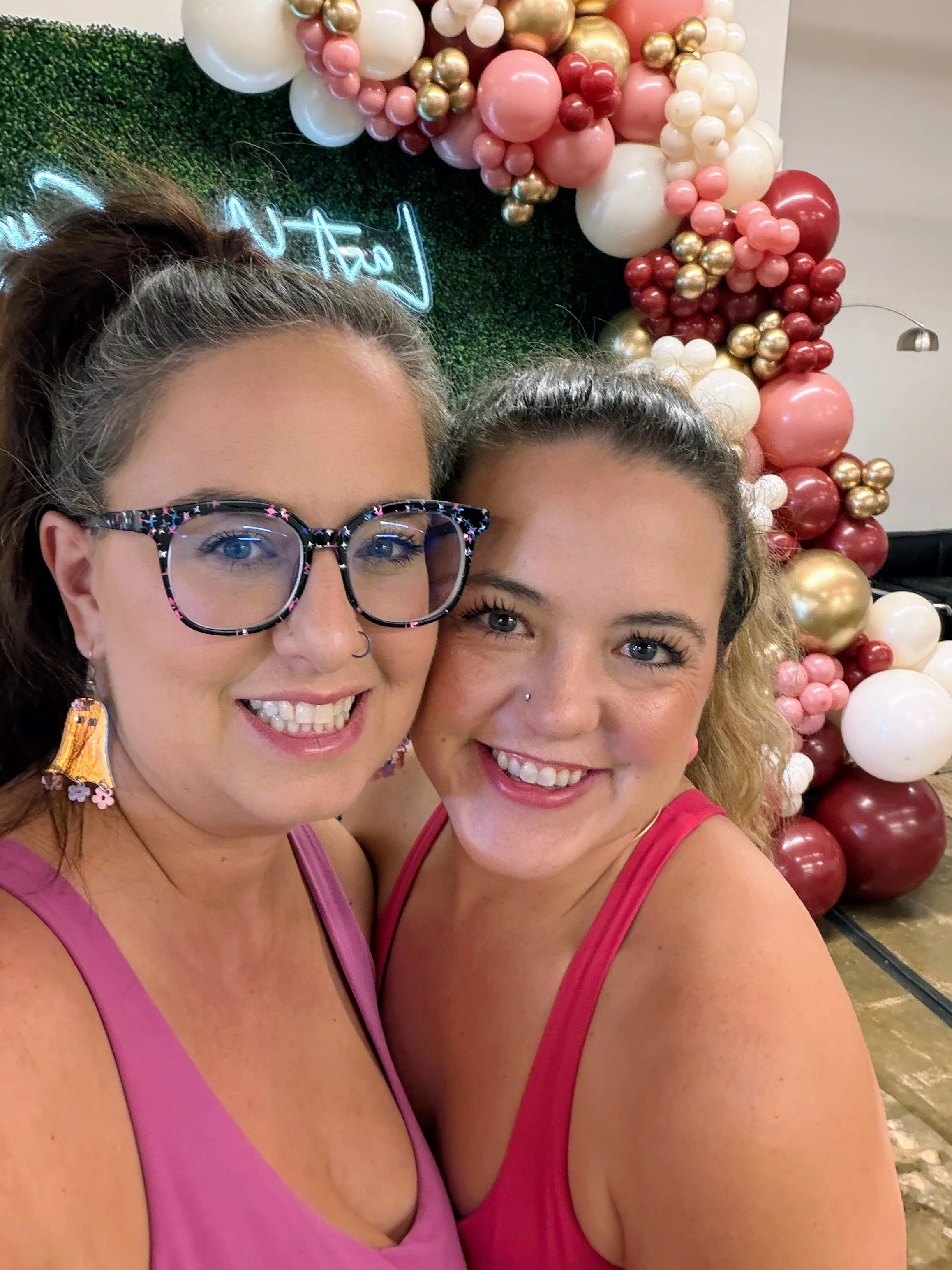 Two women smiling for a selfie in front of a decorative balloon backdrop with pink, white, red, and gold balloons. One woman has glasses and earrings, the other has a nose piercing. Both are wearing pink tops.