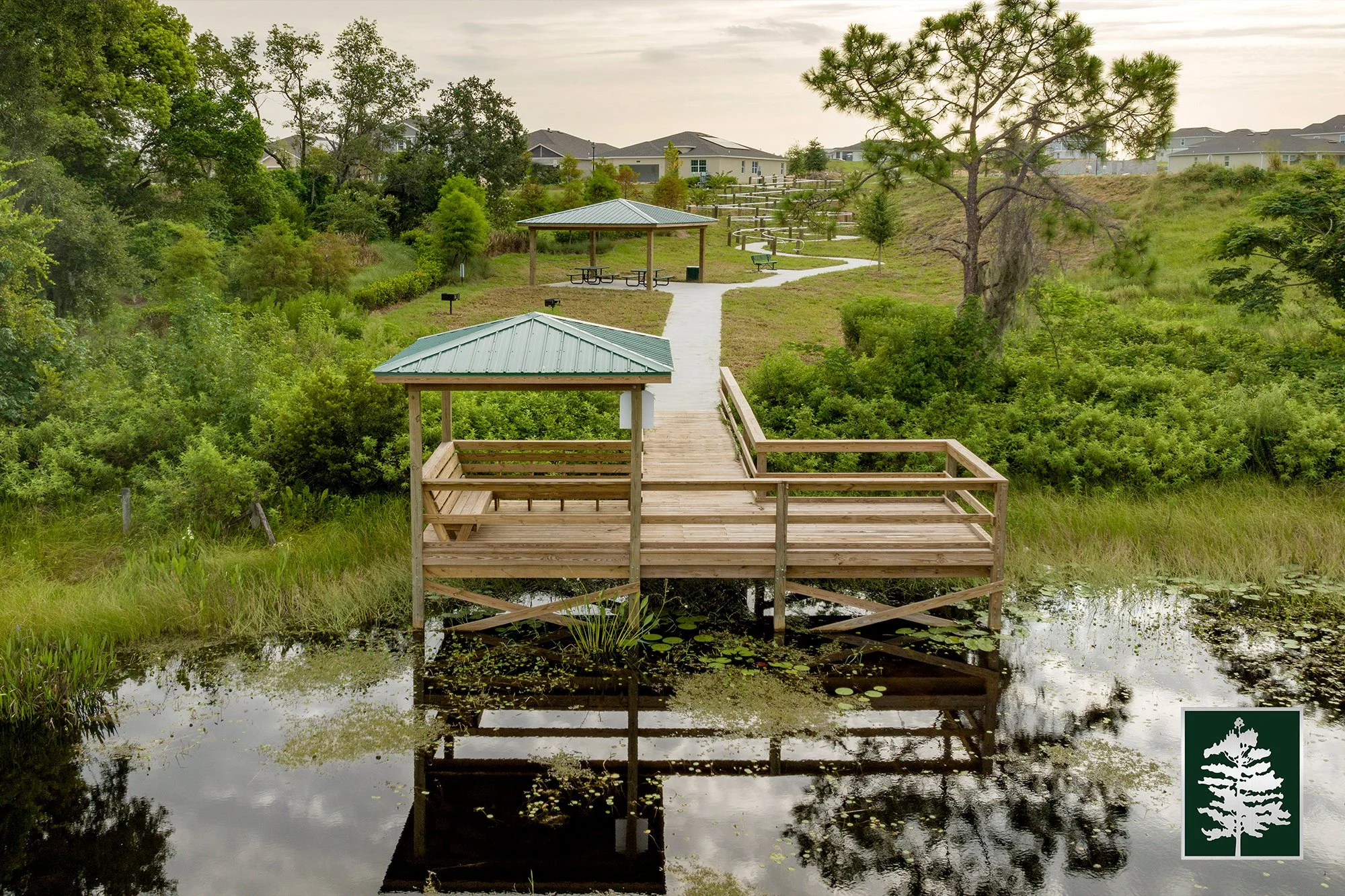 Wooden gazebo over a small pond with foliage and a walking trail leading to a park area with benches and trees, residential homes in the background.