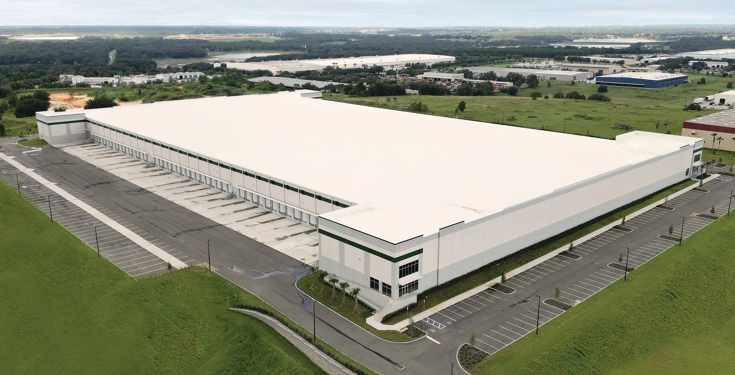 Large industrial warehouse with white roof, surrounded by empty parking lots and green grassy areas, located in an industrial park.