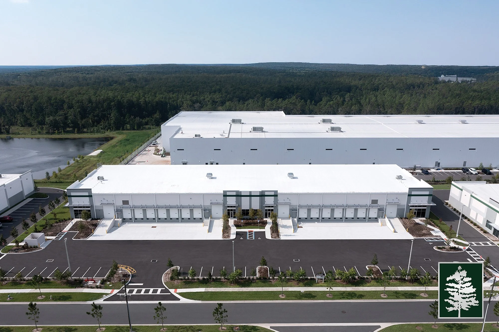 Aerial view of a modern industrial warehouse building with a parking lot and green landscaping surrounding it, located near a body of water and forested area.