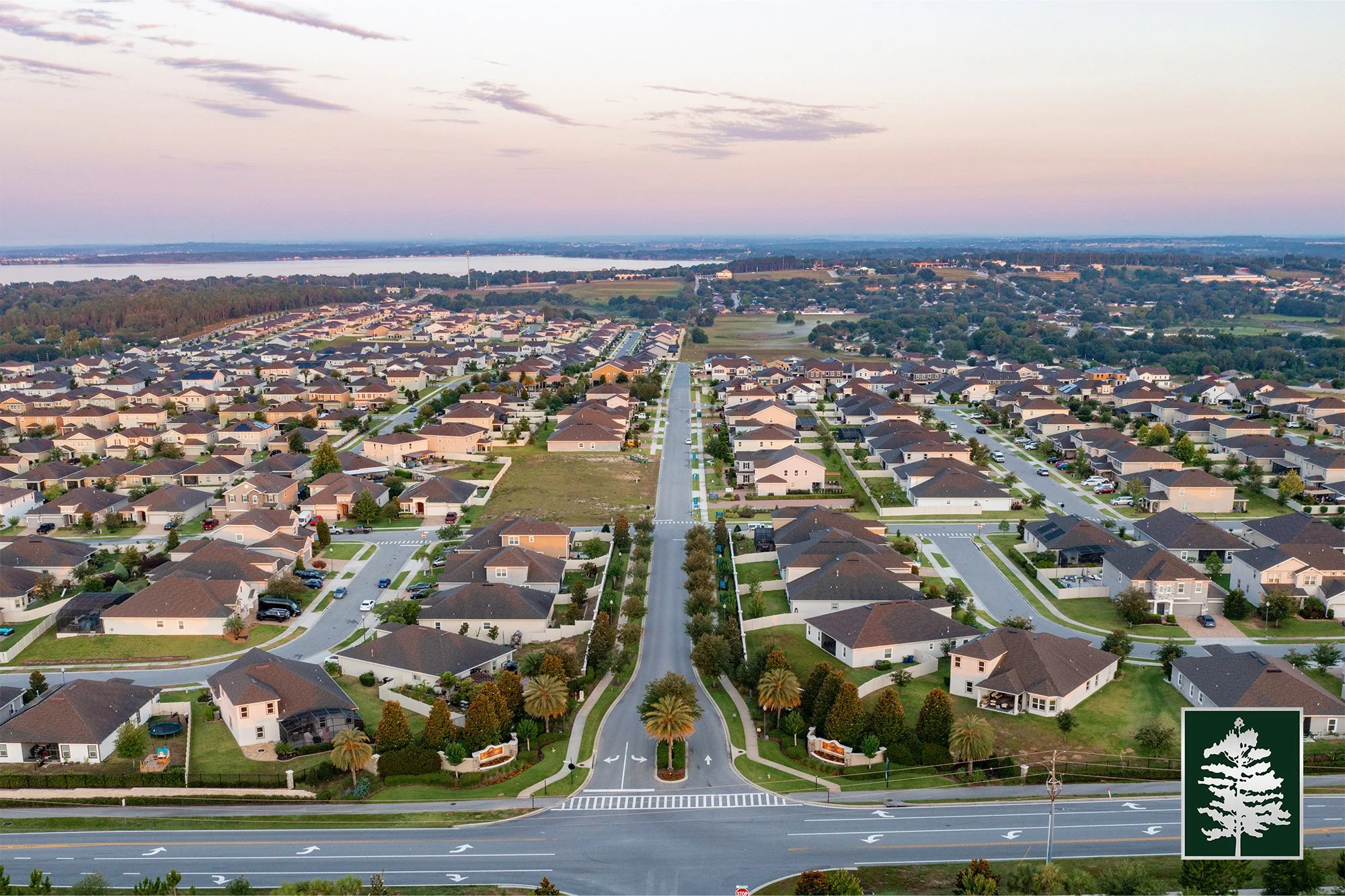 An aerial view of a suburban neighborhood with houses, streets, and green lawns, taken during sunset with a river and trees in the background.