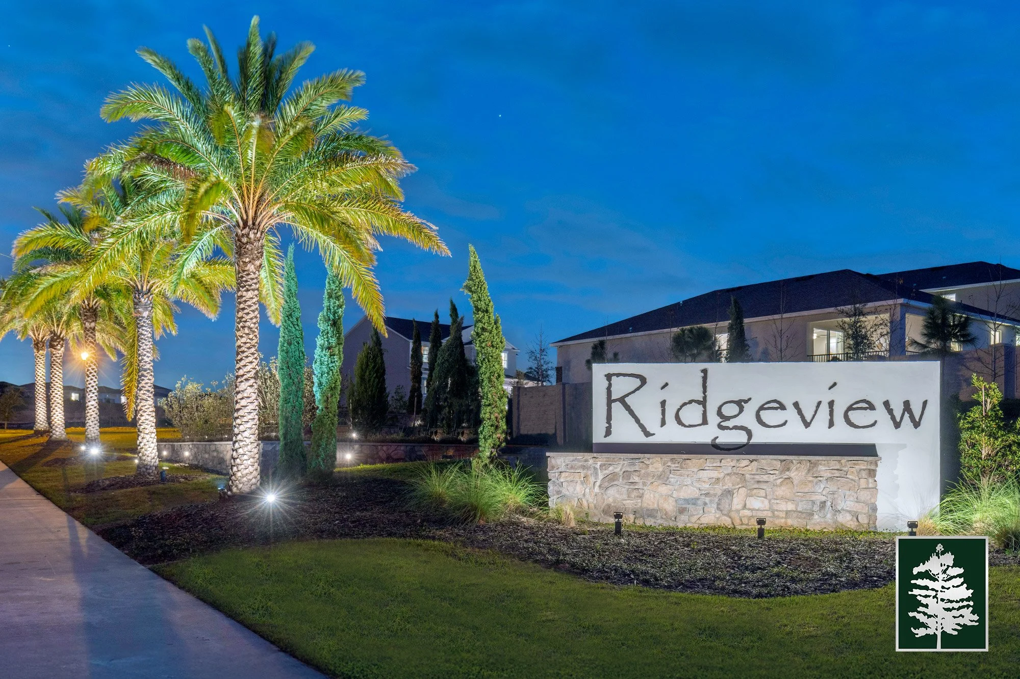 Night view of a residential community entrance with a large stone sign that reads 'Ridgeview', surrounded by palm and cypress trees, with houses in the background and decorative landscape lights.