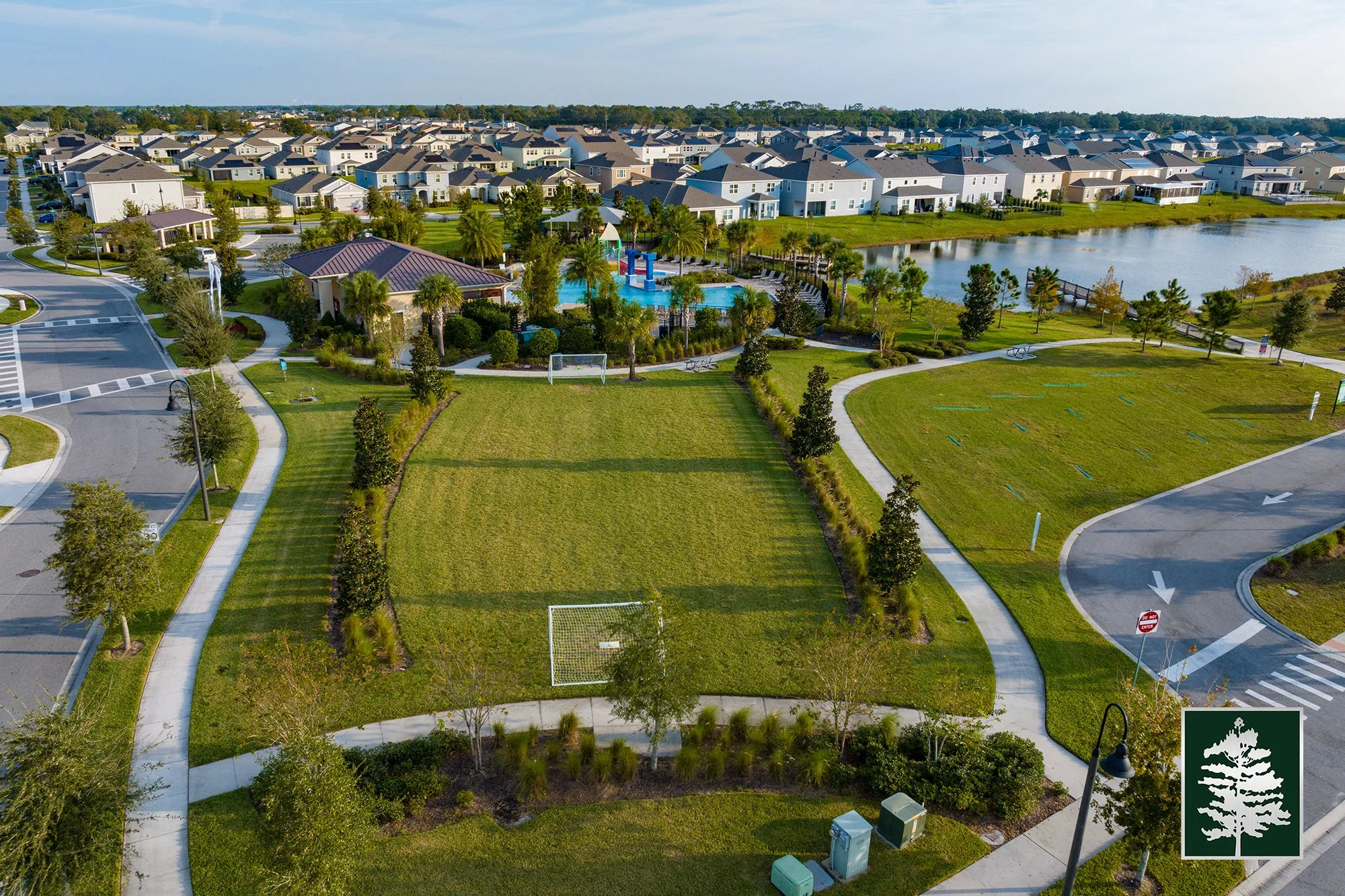 Aerial view of a suburban neighborhood with a central park area, lush greenery, walking paths, a playground, a pond, and surrounding houses.