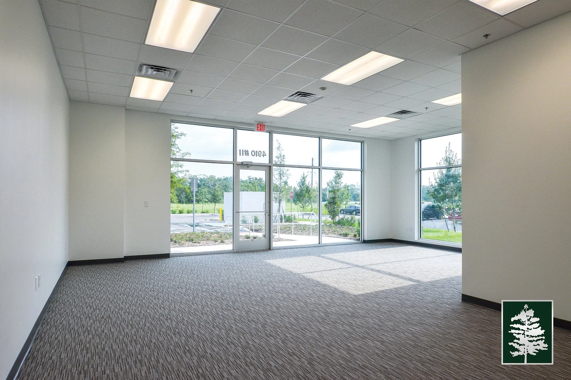 Empty commercial space with large windows and carpeted floor, outside view of parking lot and trees
