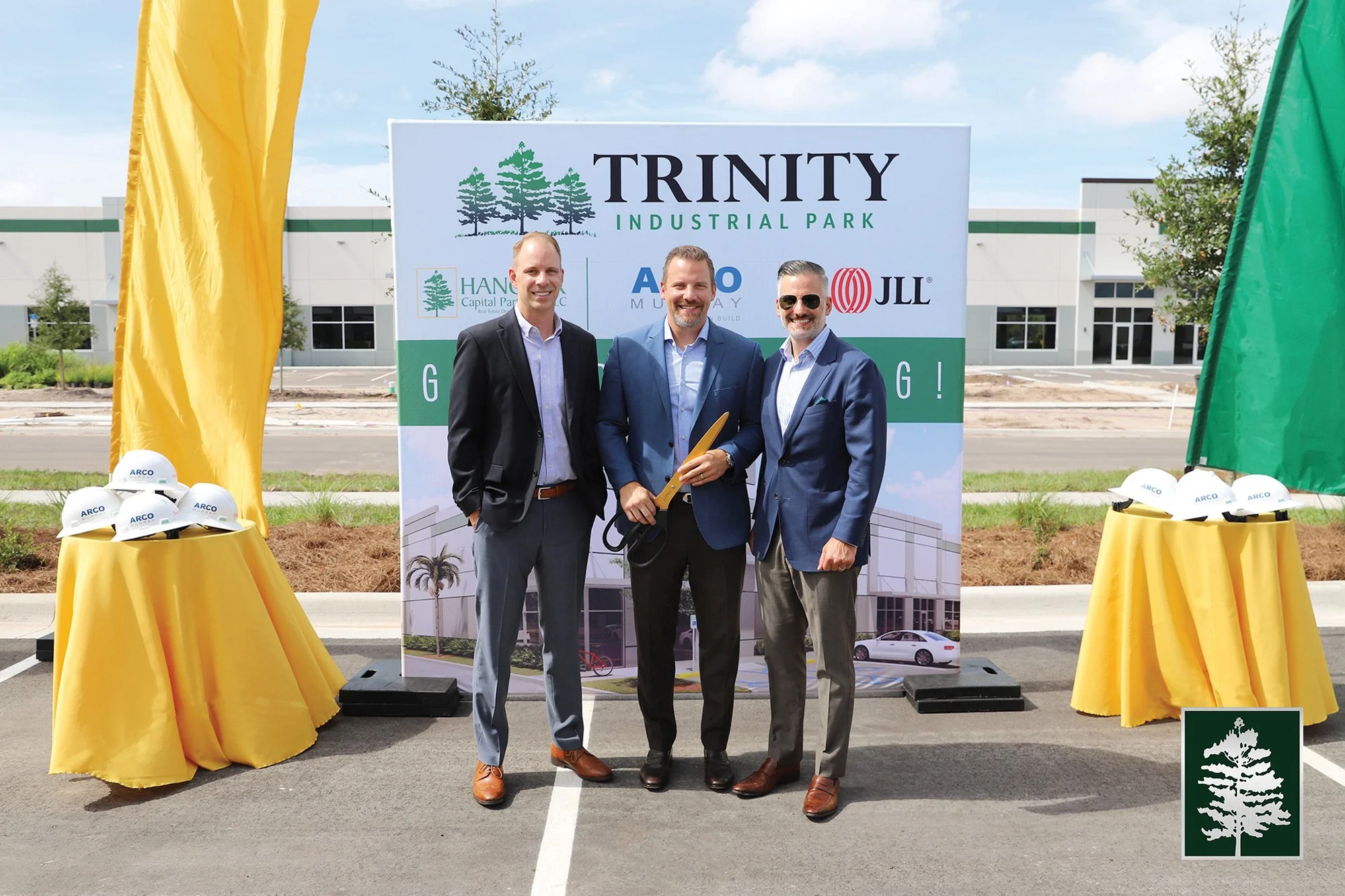Three men in suits standing in front of a sign at the groundbreaking ceremony of Trinity Industrial Park. The sign displays the Trinity logo and logos of tenants Hand Capital Partners, A M Murray, and JLL. Two tables with yellow tablecloths and white hard hats with the ARCO logo are on either side of the men. The background shows an under-construction industrial building and a partly cloudy sky.