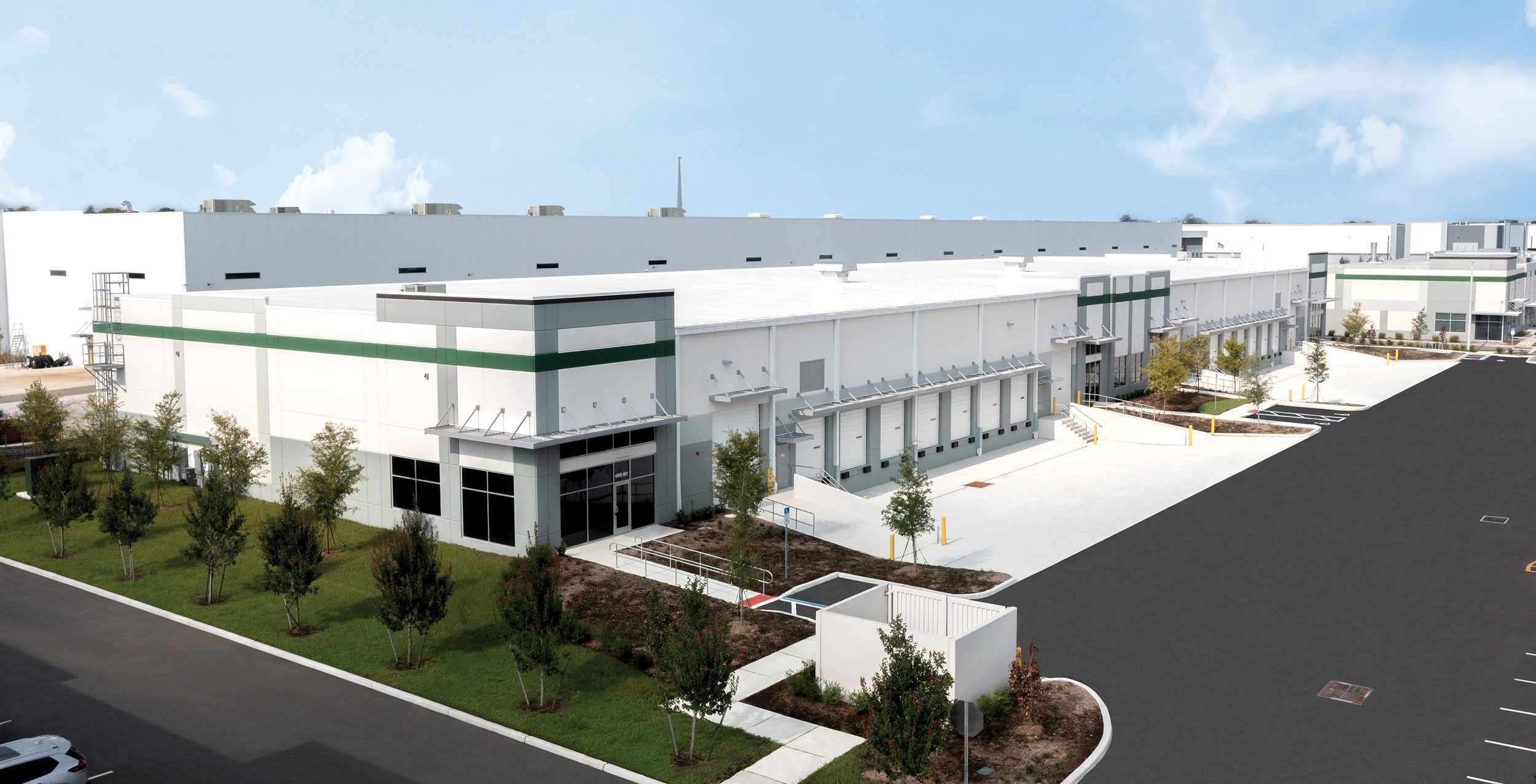A large modern warehouse with white walls and green accents, multiple loading docks, and surrounding parking area with trees and landscaping under a partly cloudy sky.