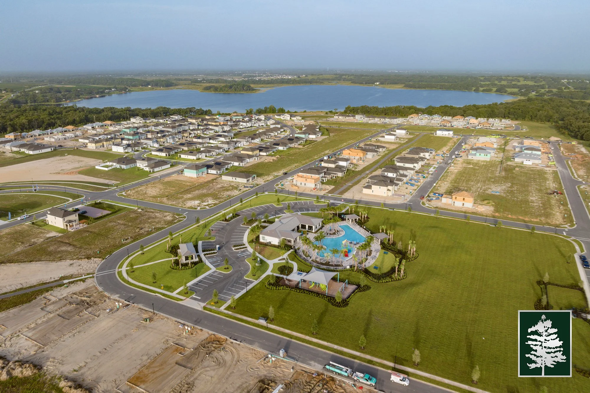 Aerial view of a residential community under construction near a lake, featuring houses in development, roads, a communal pool area with a playground, and open grassy spaces.