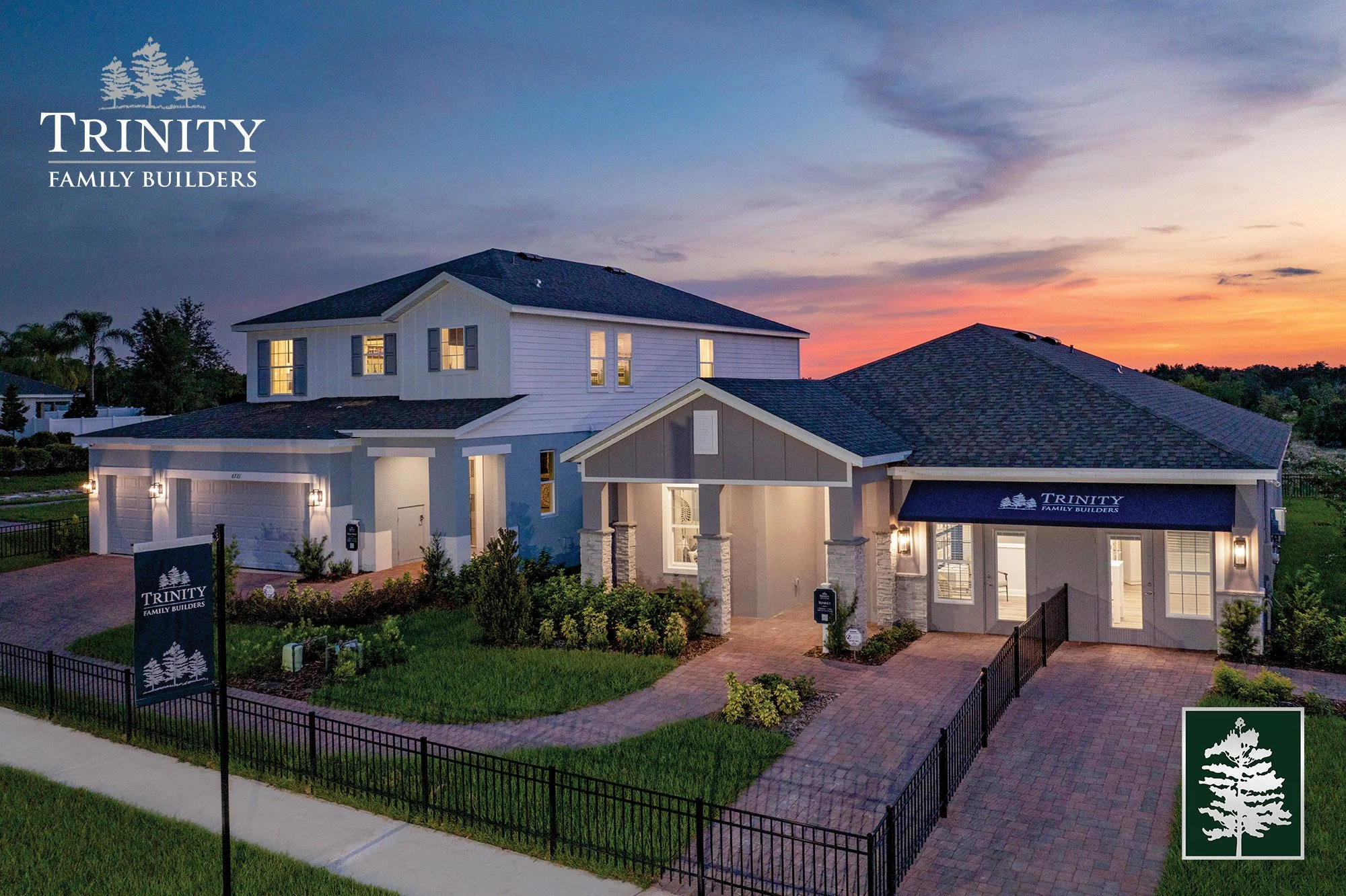 Newly built two-story house with a front yard, driveway, and landscaping, illuminated at dusk with lights inside and outside, sign and awning display 'Trinity Family Builders', and a logo in the bottom right corner